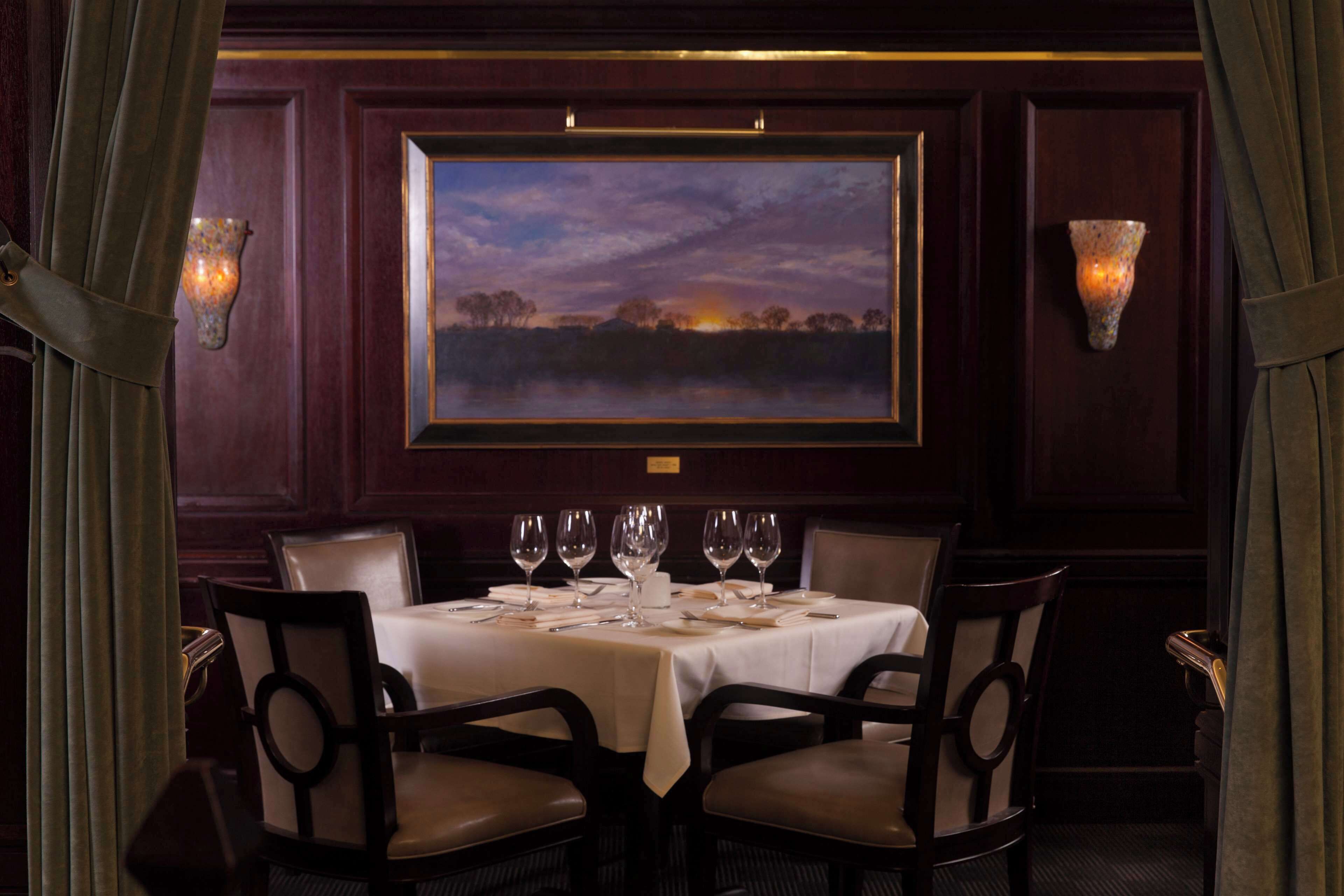 A dining area with a table set for six, flanked by two wall-mounted light fixtures and a framed landscape painting above.