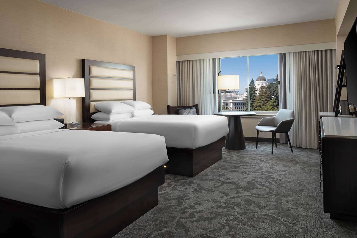 A hotel room with two beds, a desk, a chair, and a view of a cityscape.