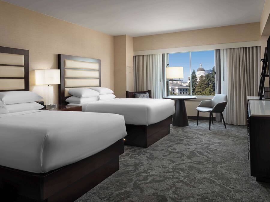 A hotel room with two beds, a desk, a chair, and a view of a cityscape.