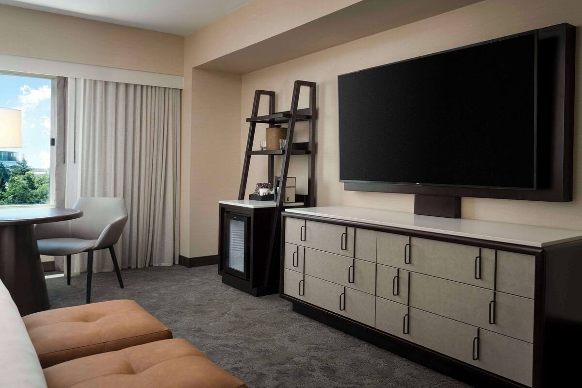 A hotel room with a large flat-screen TV mounted on the wall, a small desk with a chair, a sofa, and a minibar.