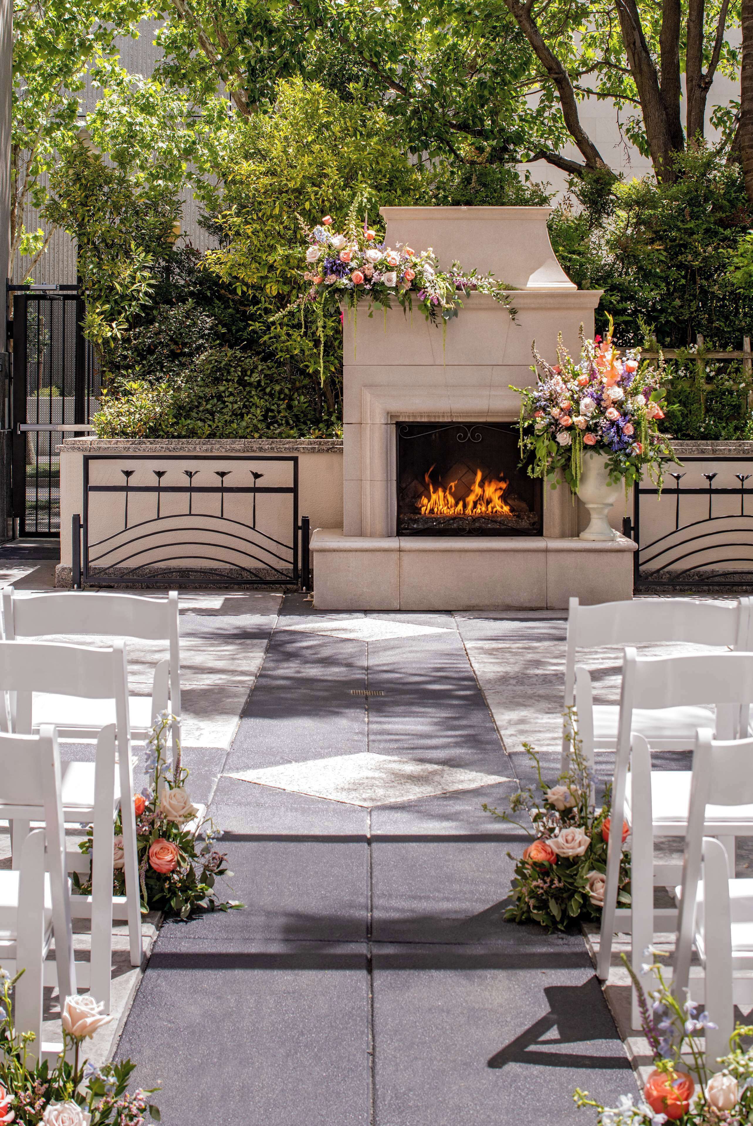 An outdoor wedding aisle with white chairs, a stone fireplace with a fire, and floral arrangements.