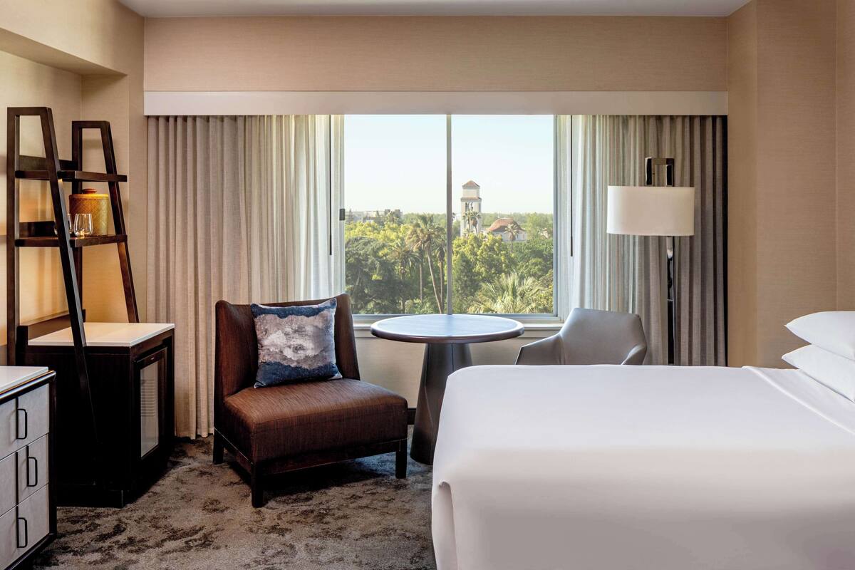 A hotel room with a bed, a chair, a small table, a desk, and a lamp.