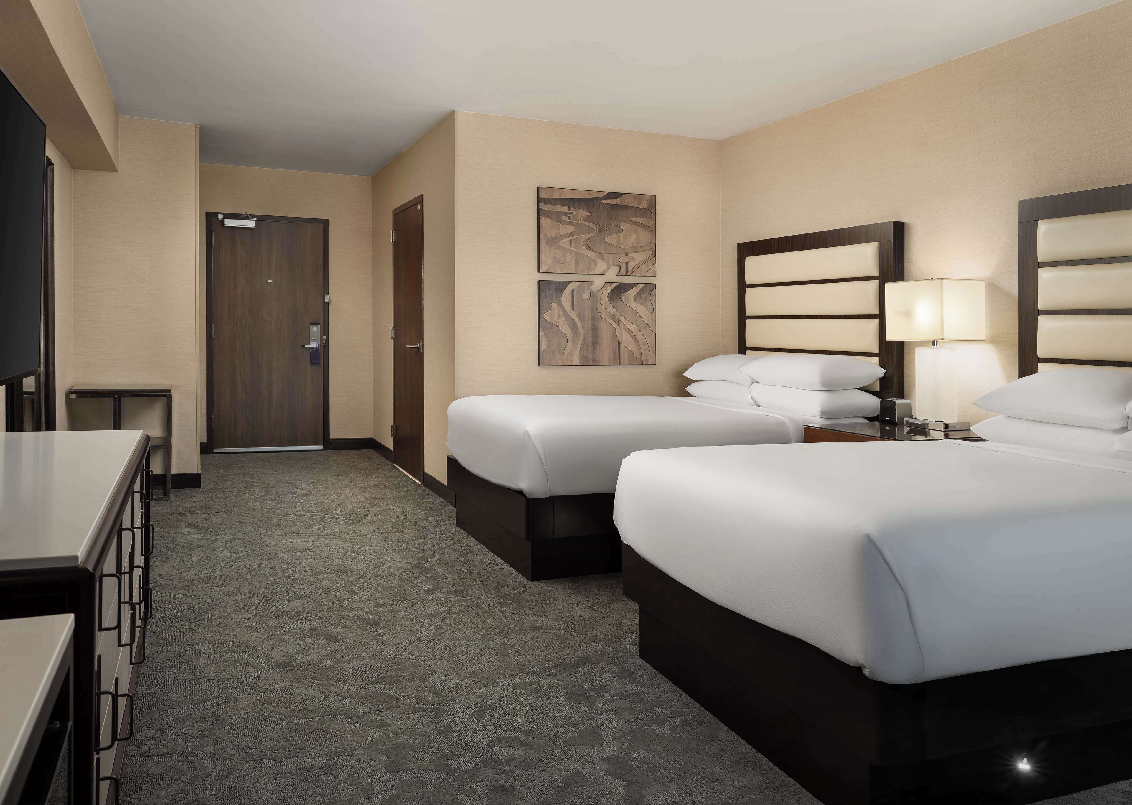 Quarto de hotel com duas camas, uma escrivaninha, uma cadeira, uma porta e uma obra de arte na parede.