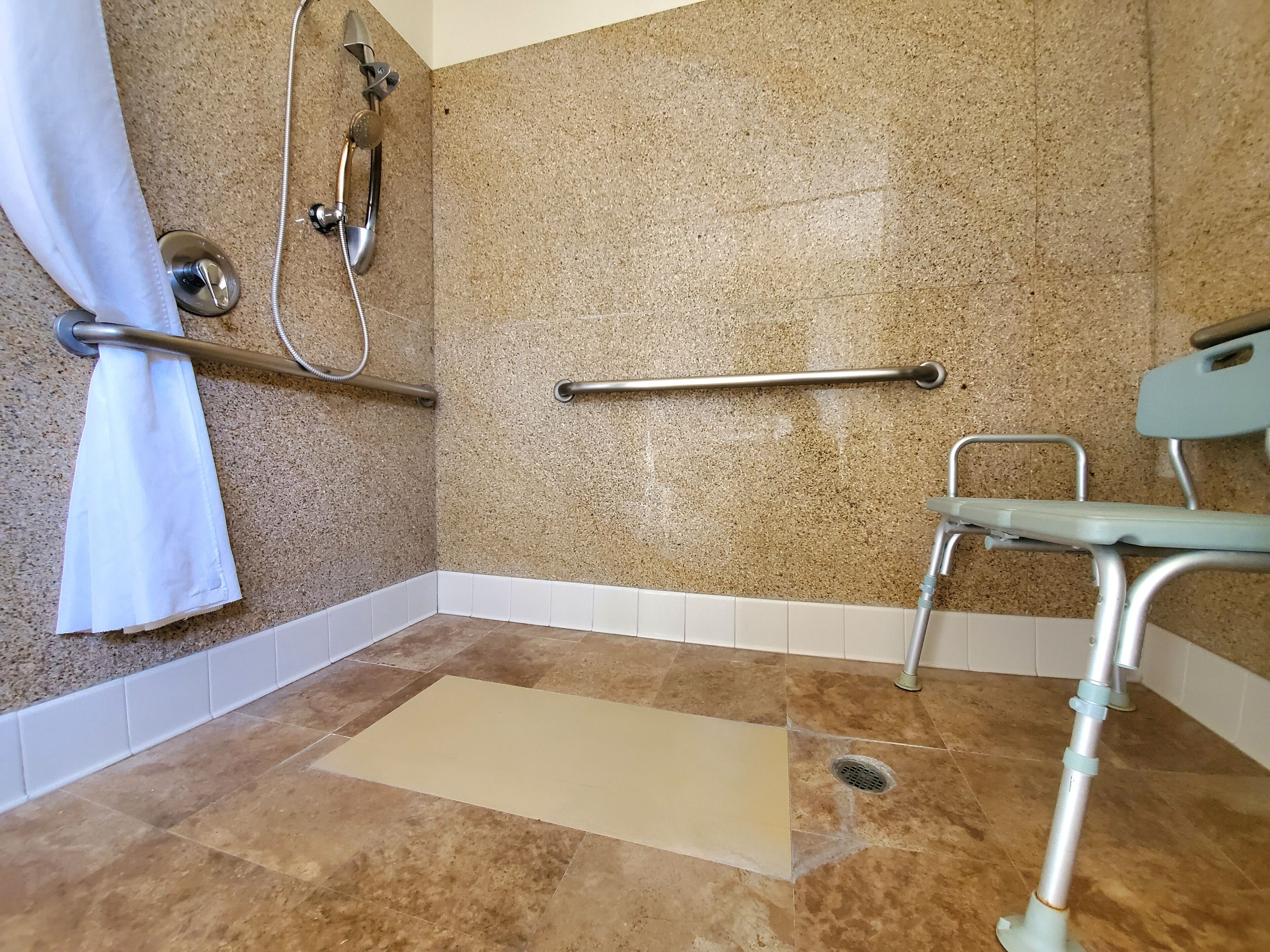 A bathroom with a shower, grab bars, a wheelchair-accessible bench, and a white curtain.