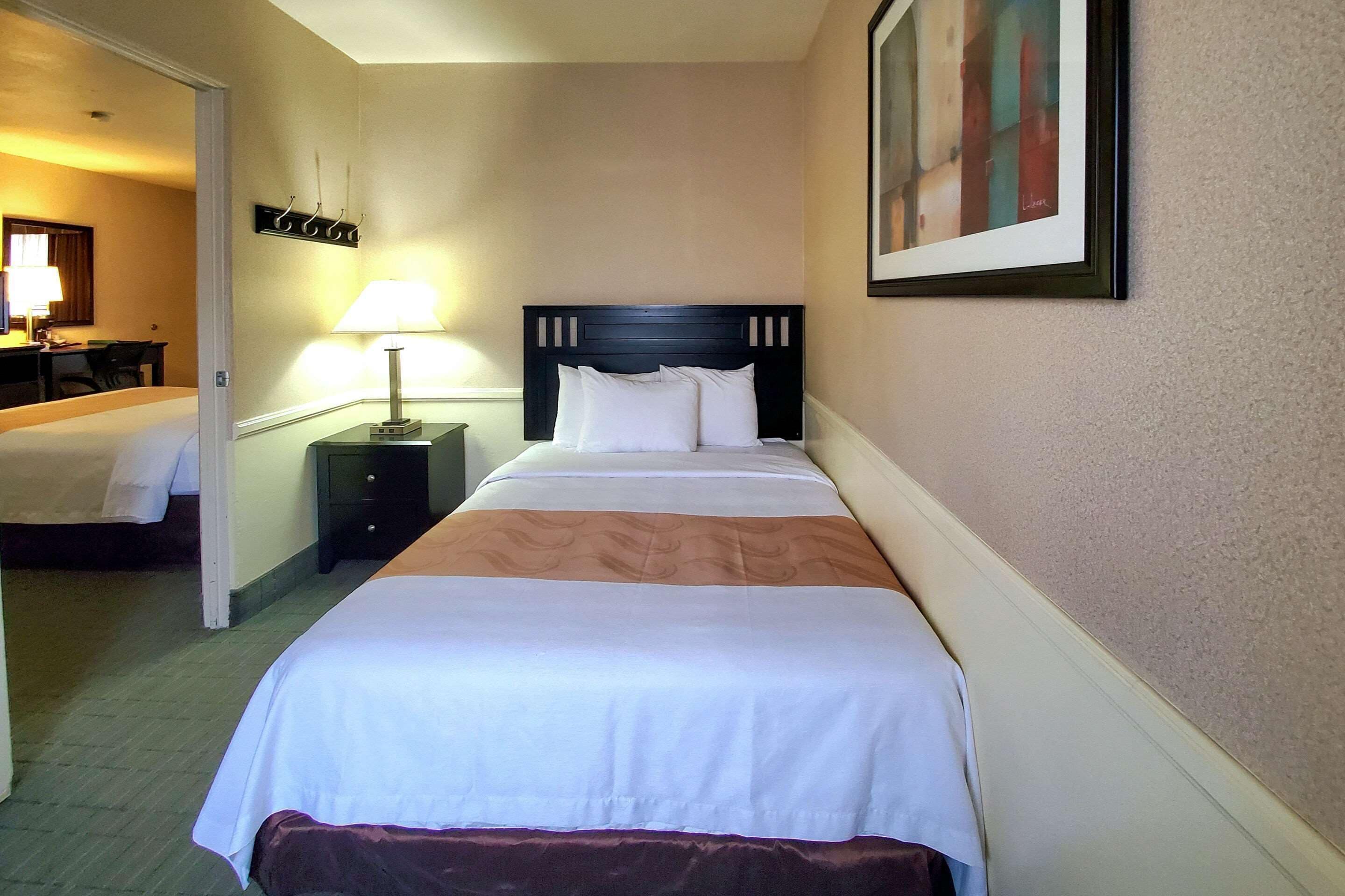 A hotel room with two beds, a bedside table with a lamp, a framed picture on the wall, and a desk with a chair in the background.