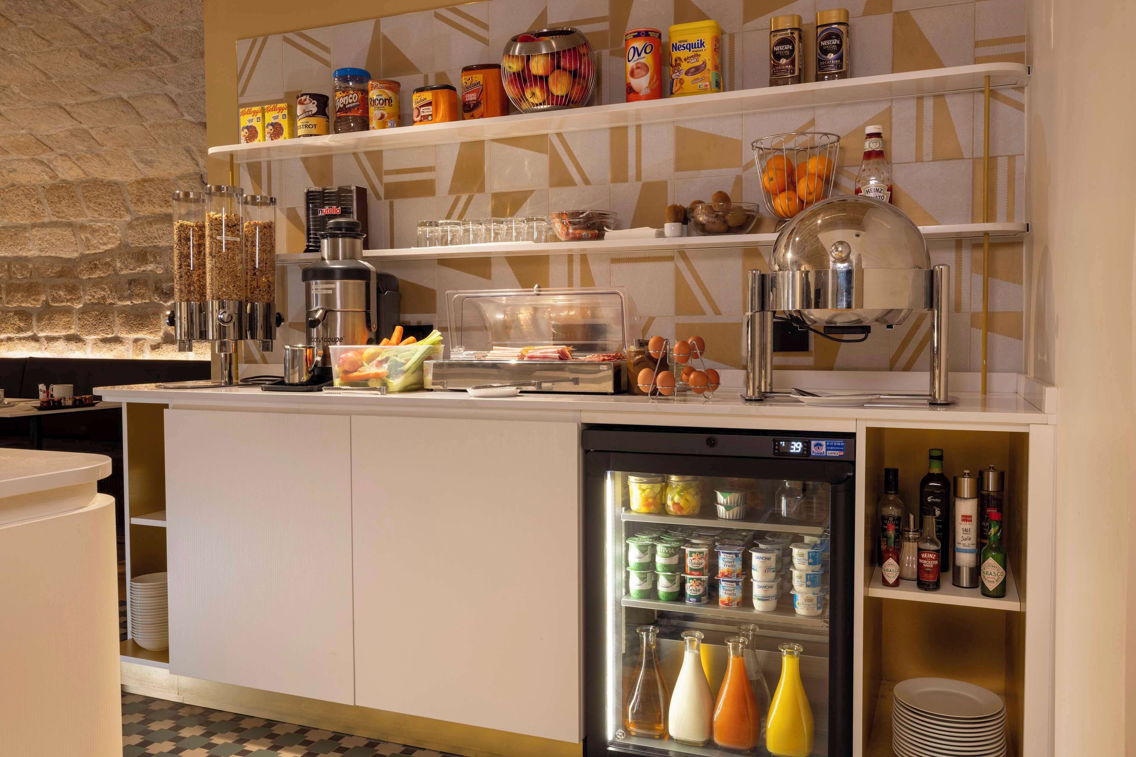 Una barra de desayuno con cafetera, cereales y salsas. Hay un refrigerador con bebidas y una repisa con condimentos.