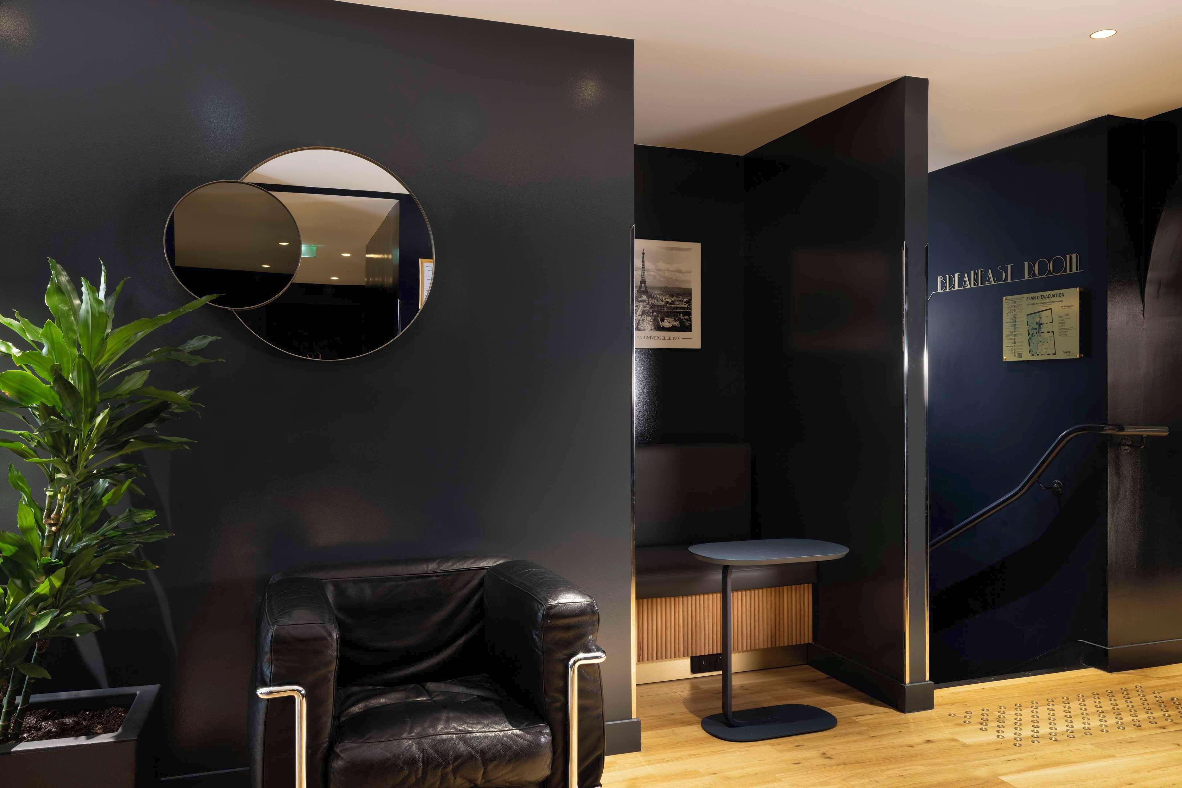 A modern hotel room with a dark wall, a round mirror, a leather chair, a small round table, and a staircase leading to the breakfast area.
