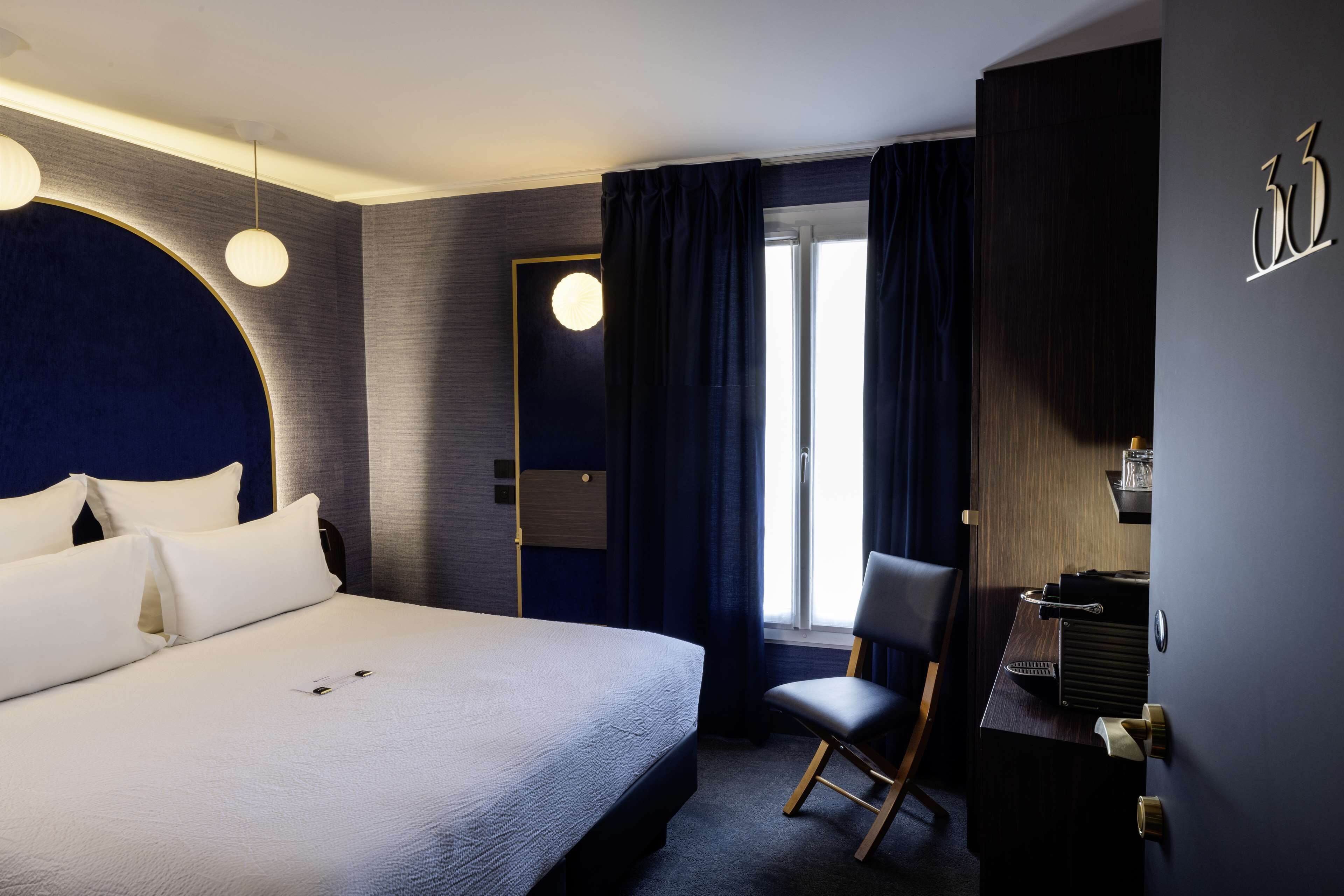 A modern hotel room with a large bed, a desk with a chair, a round light fixture, and a view of the outdoors through a window.
