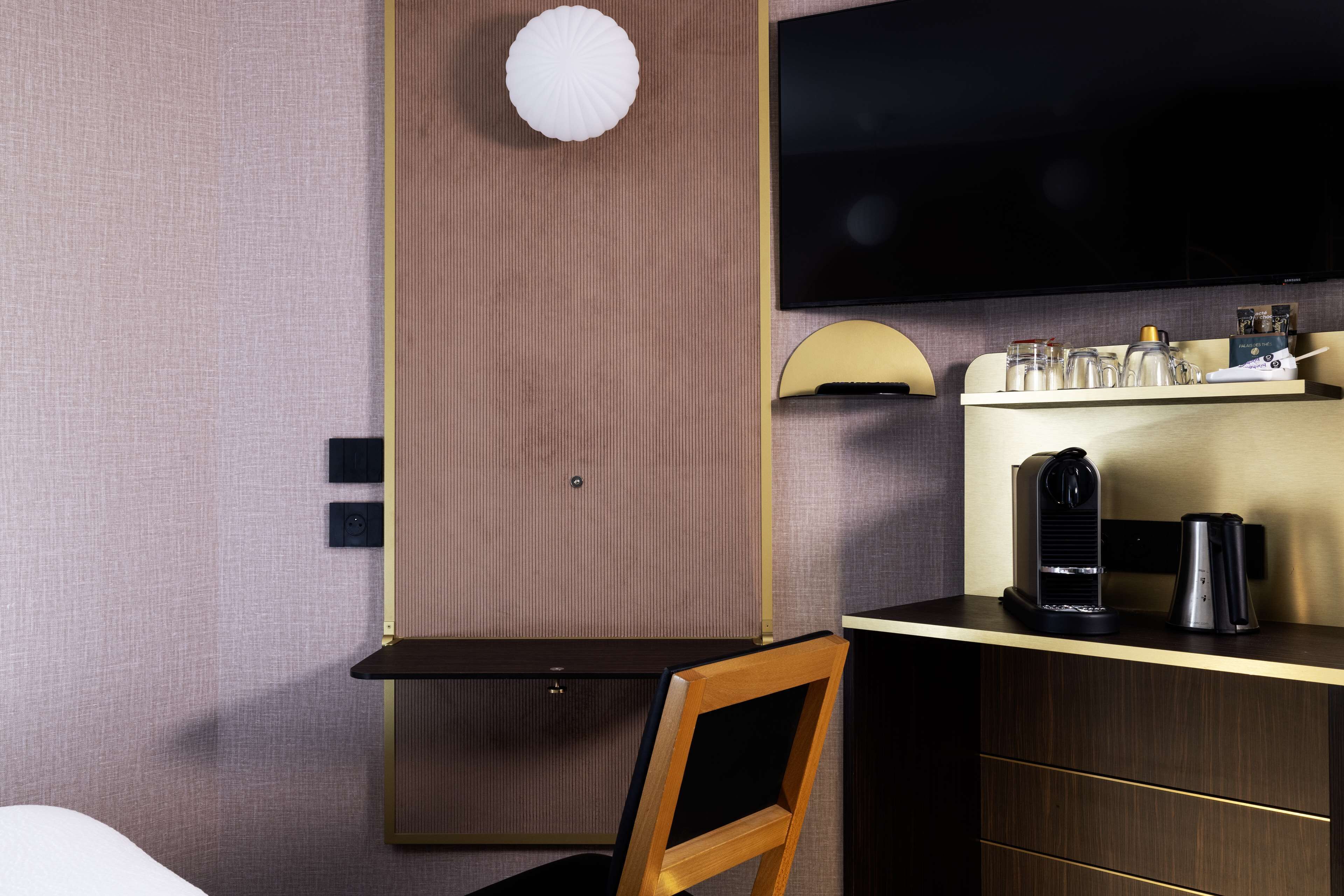 Une chambre d’hôtel comprenant un lit, un bureau avec une chaise, une télévision, une machine à café et une étagère contenant divers objets.