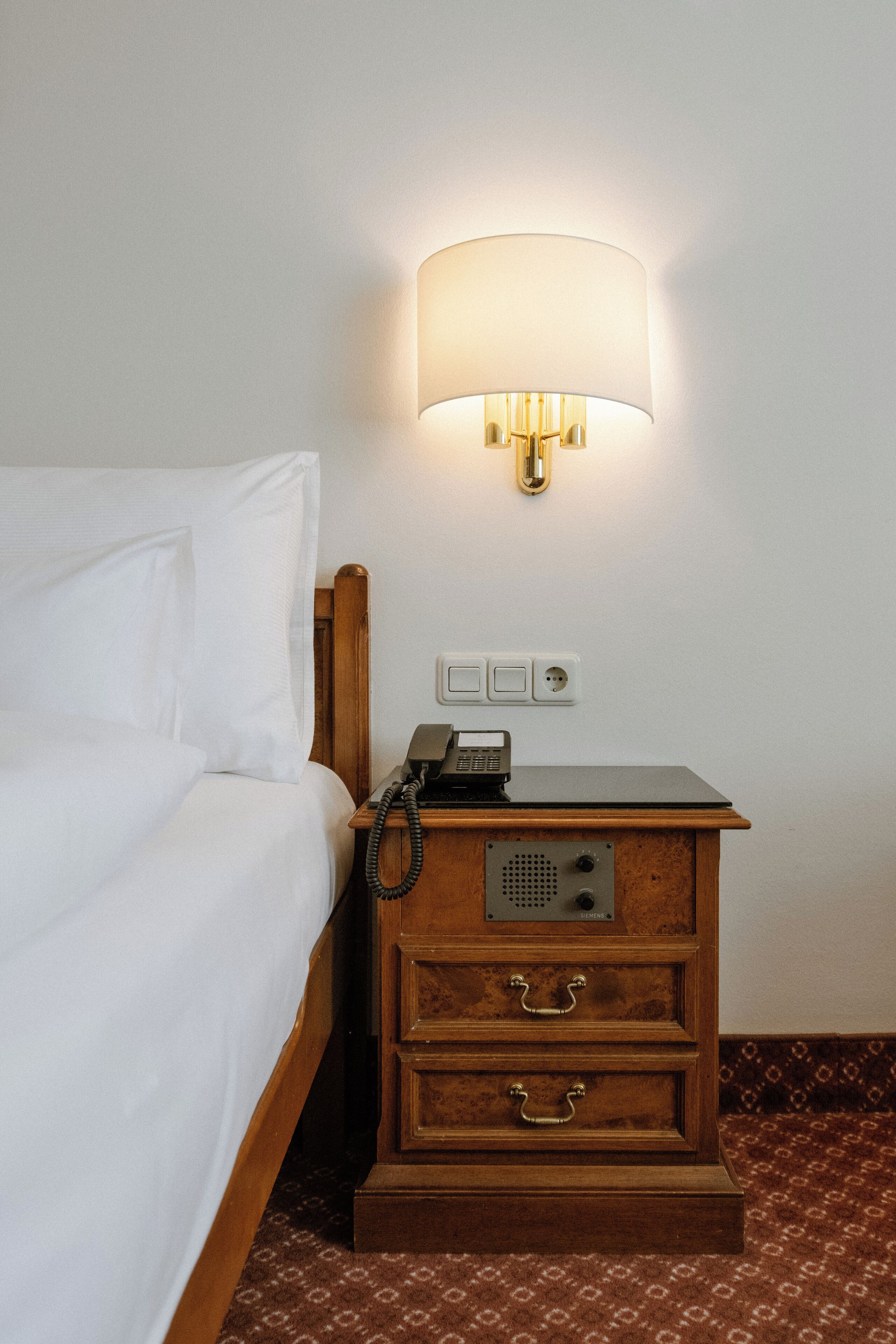 A hotel room with a bed, a wooden nightstand with a lamp, a telephone, and a power outlet.