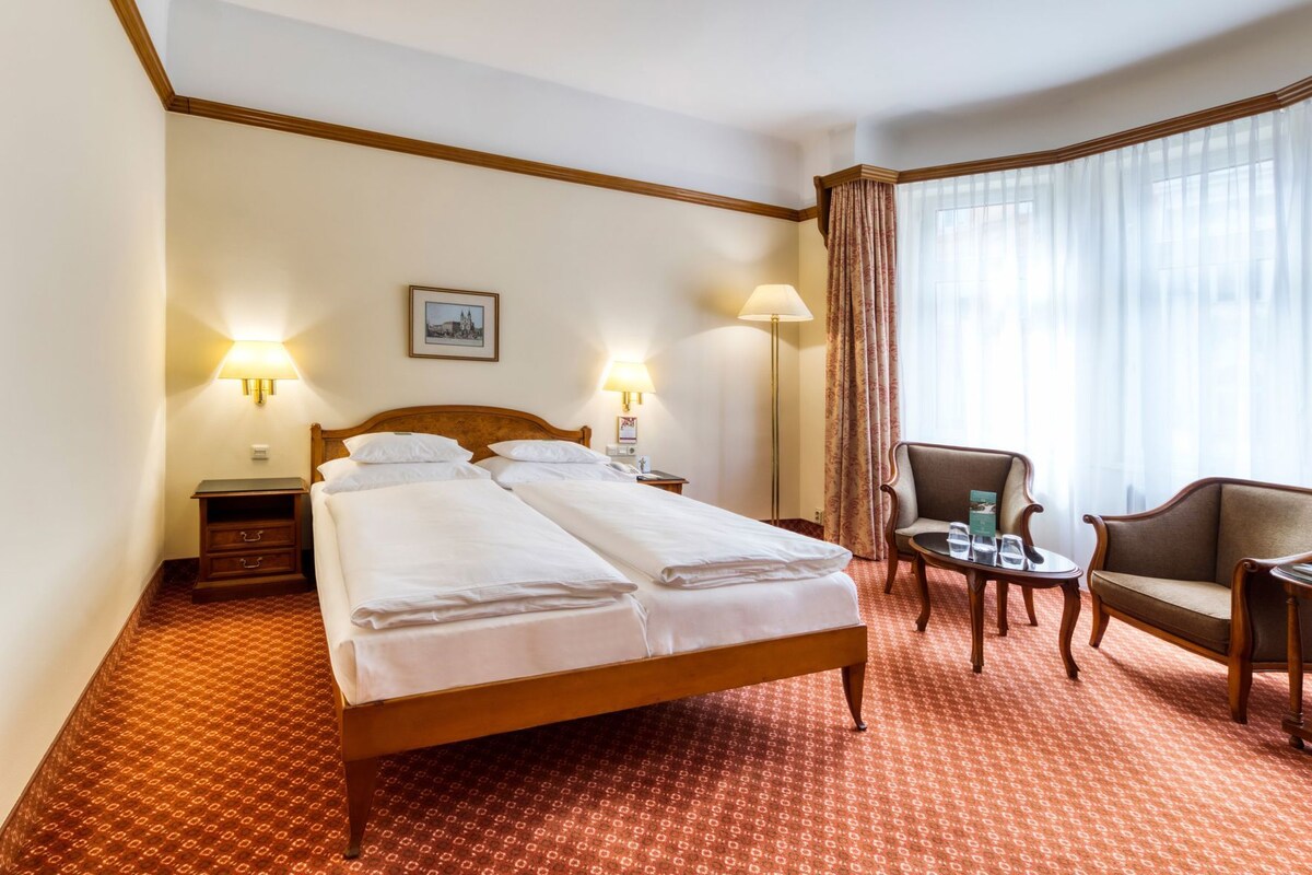 A hotel room with a double bed, two bedside tables, a chair, a small table, and a window with curtains.