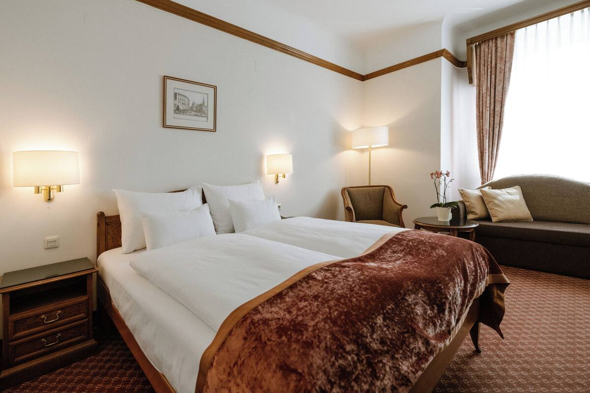 A hotel room with a large bed, two bedside lamps, a sofa, a small table with a plant, and a framed picture on the wall.