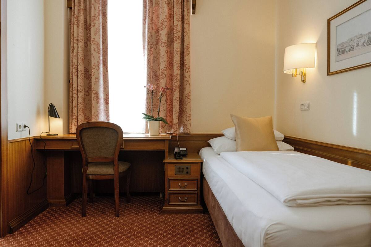 A hotel room with a wooden desk, a chair, a bed with white linens, a nightstand with a lamp, and a window with patterned curtains.