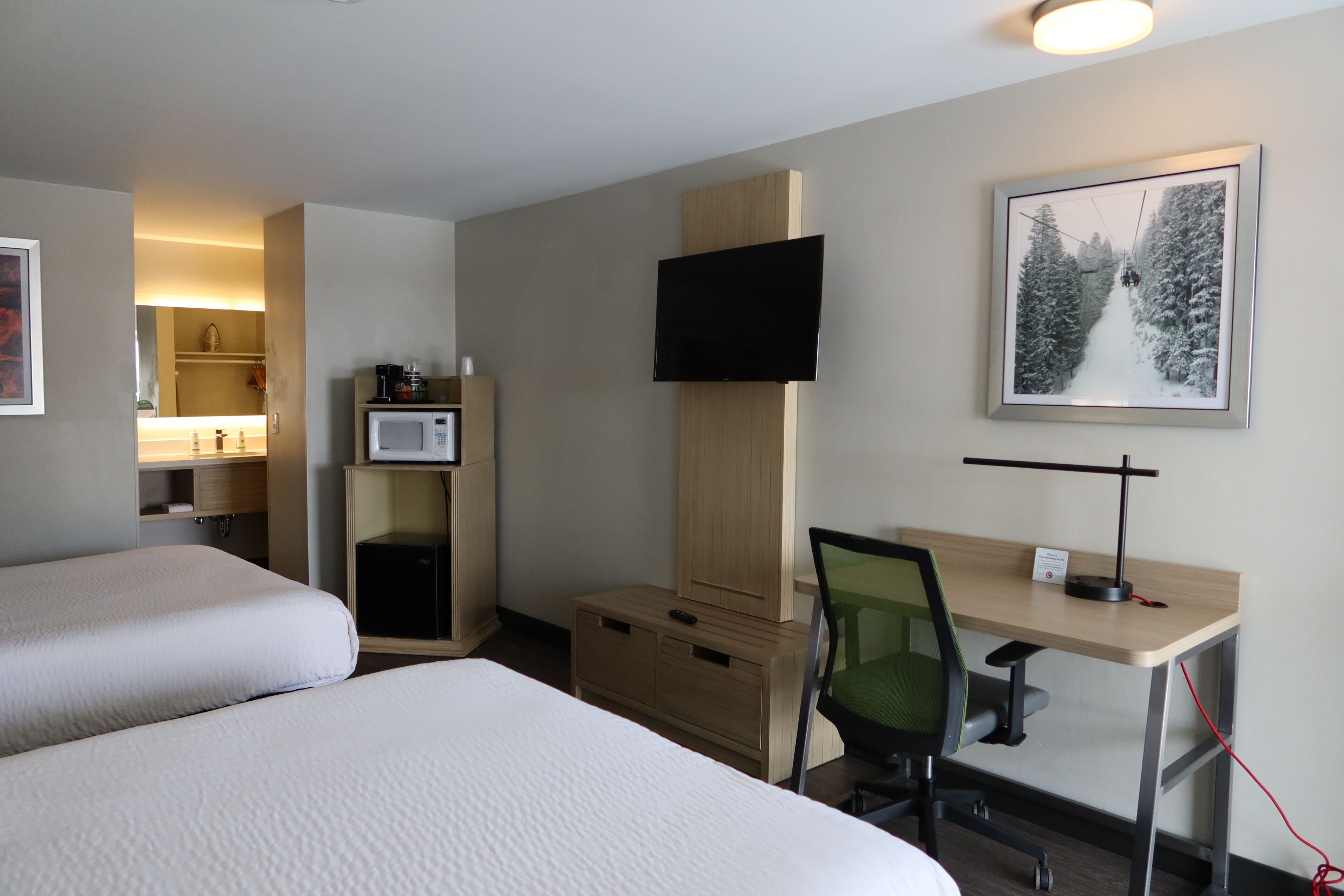 Habitación de hotel con dos camas, un escritorio con una silla, un televisor montado en la pared, un microondas y un cuadro enmarcado de un paisaje nevado.