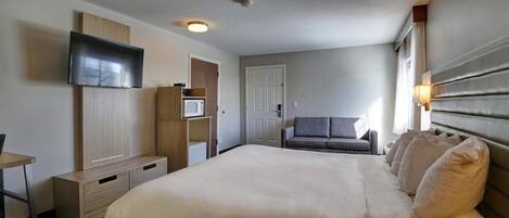 A hotel room with a large bed, a TV mounted on the wall, a desk with a chair, a sofa, and a door leading to another room.