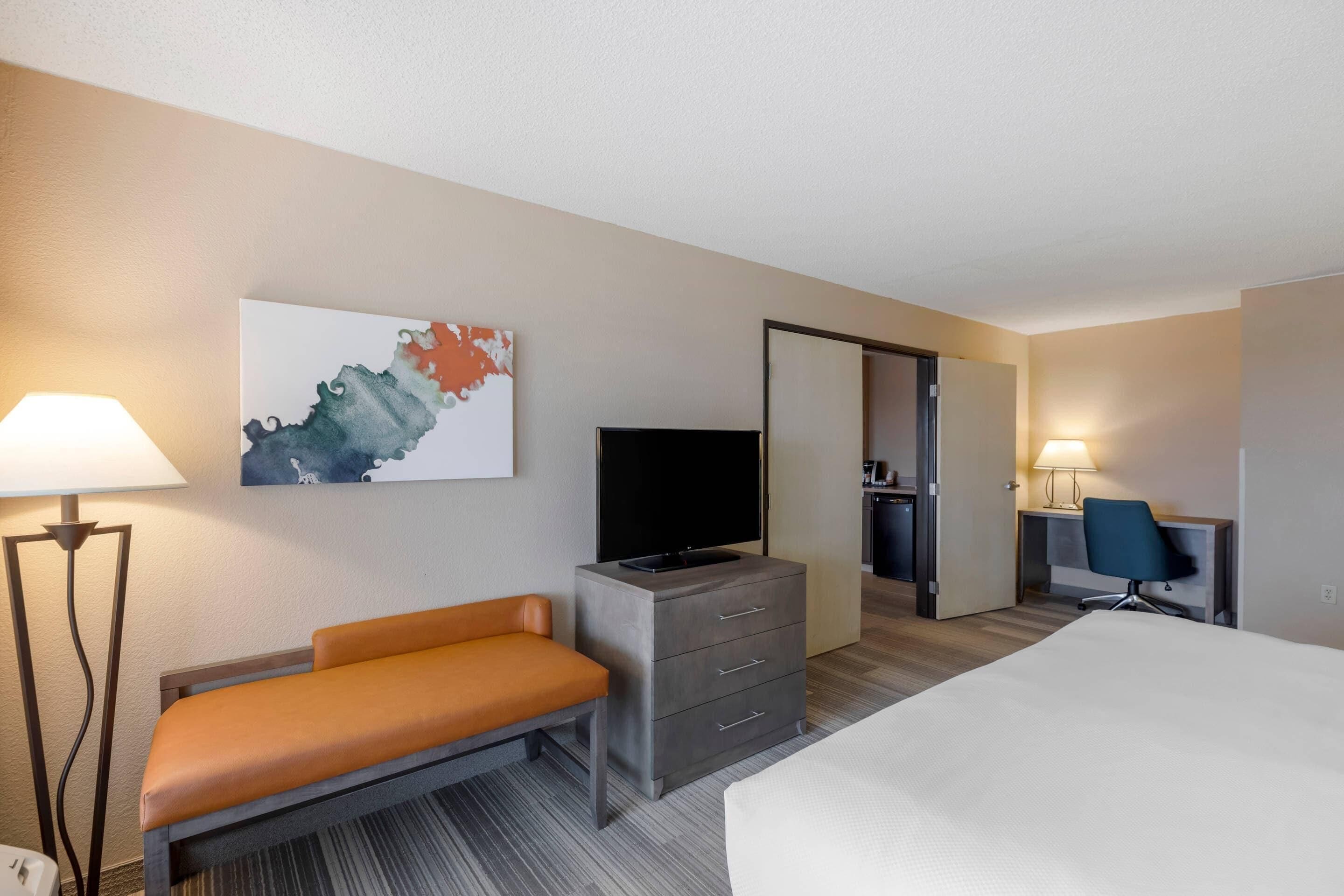 A hotel room with a bed, a desk with a chair, a television, a dresser, and a lamp.