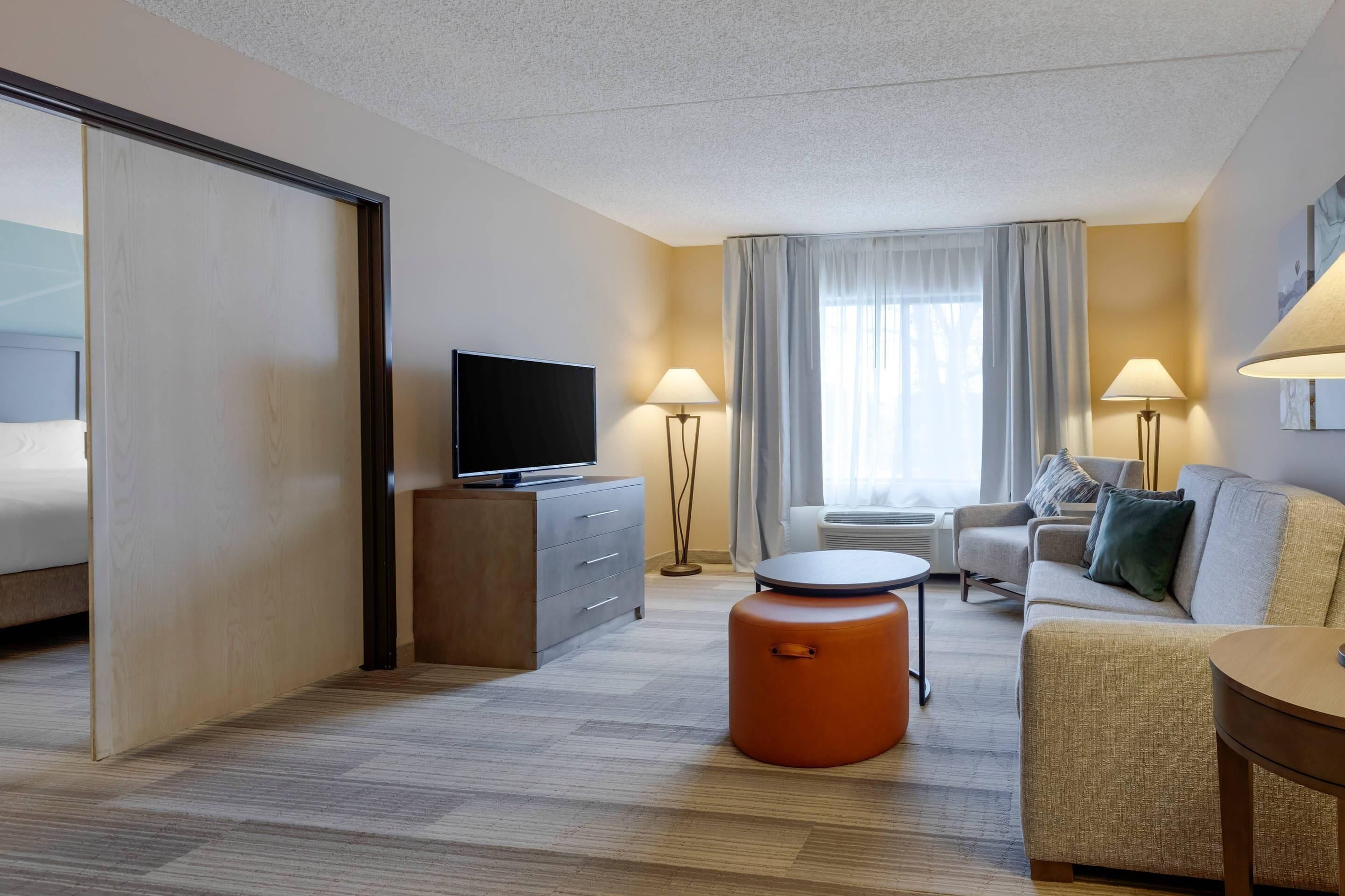 A hotel room with a bed, a television, a sofa, a coffee table, and a small orange ottoman.