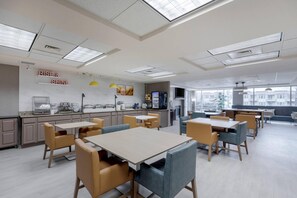 A modern dining area with tables and chairs, a kitchen counter with appliances, and a large window offering a city view.