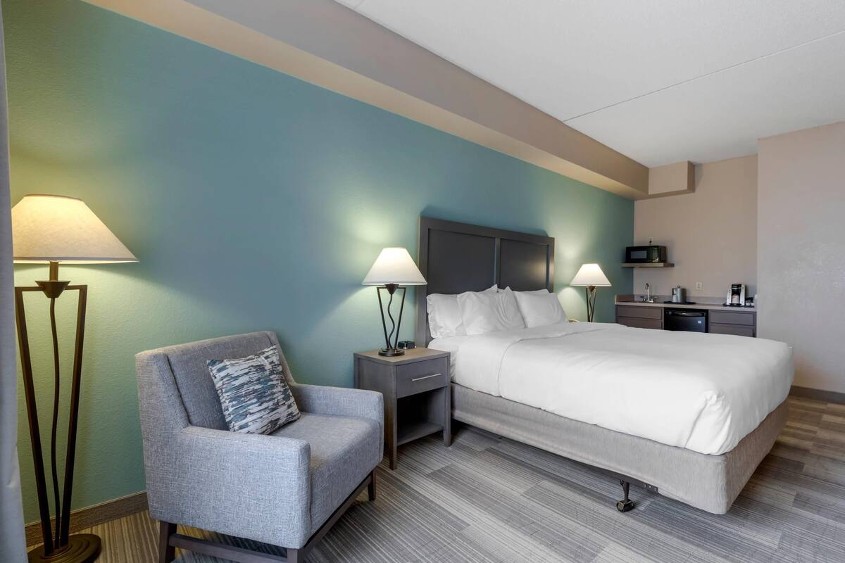 A hotel room with a bed, a chair, a nightstand with lamps, a microwave, and a small kitchenette.