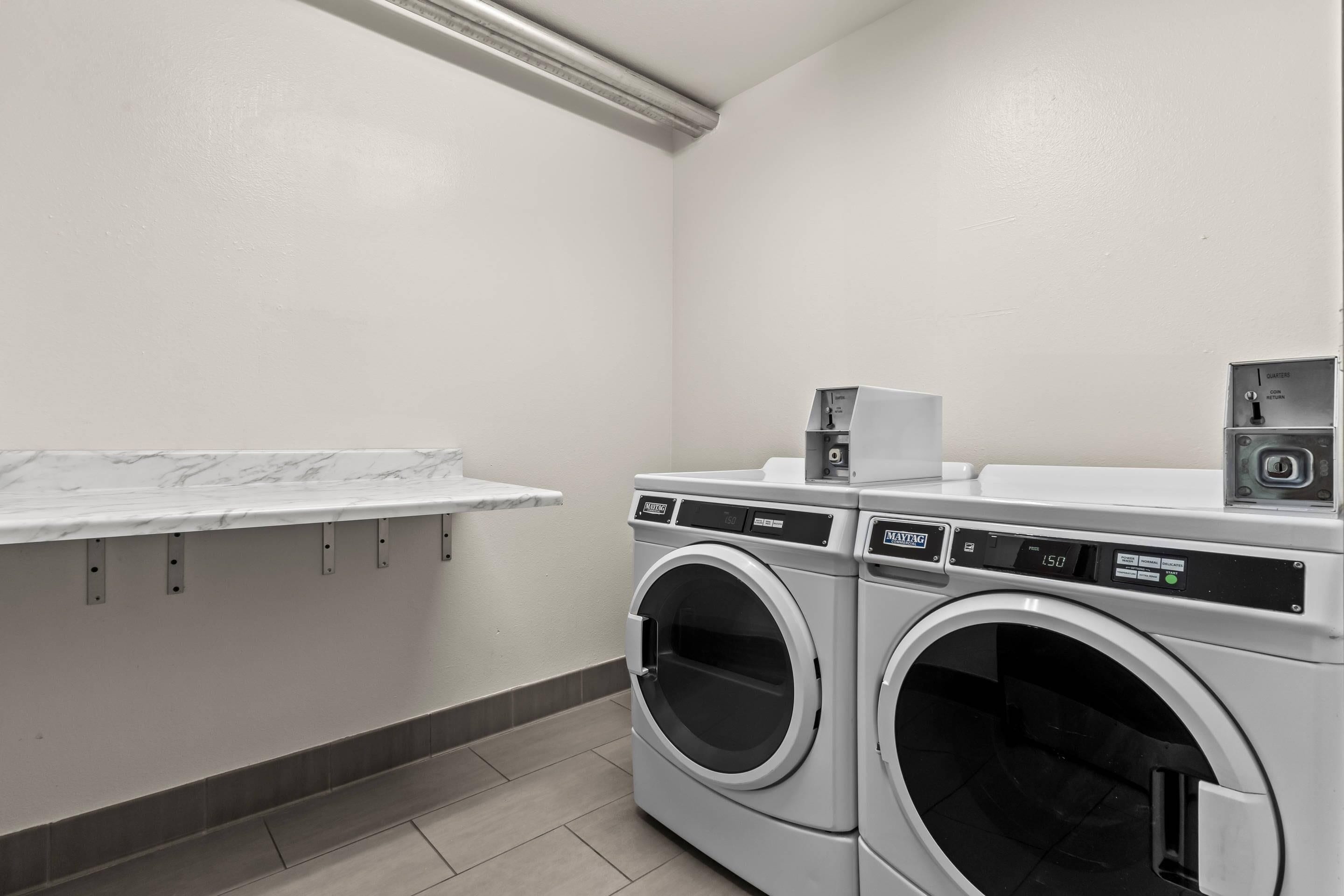 A laundry room with two front-loading washing machines, a drying machine, and a small shelf with a white marble countertop.