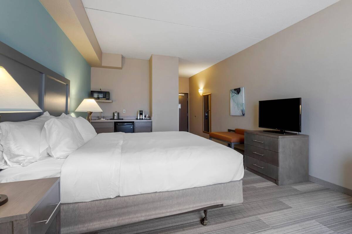 A hotel room with a large bed, a desk with a lamp, a television, and a small kitchenette.