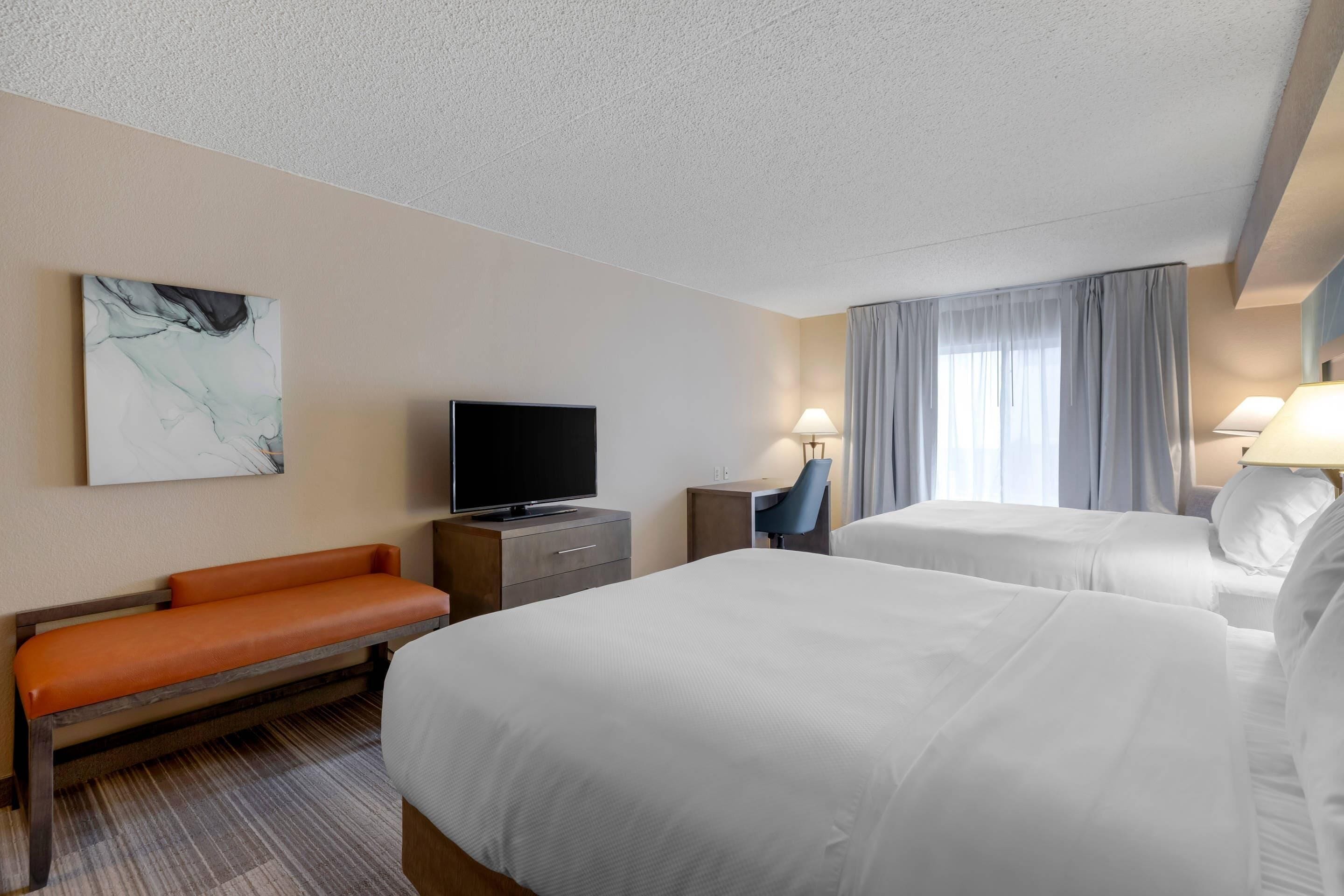 A hotel room with two beds, a desk, a television, a bench, and a painting on the wall.