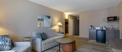 A modern hotel room with a grey sofa, a round coffee table, and a kitchenette area with a microwave and refrigerator.