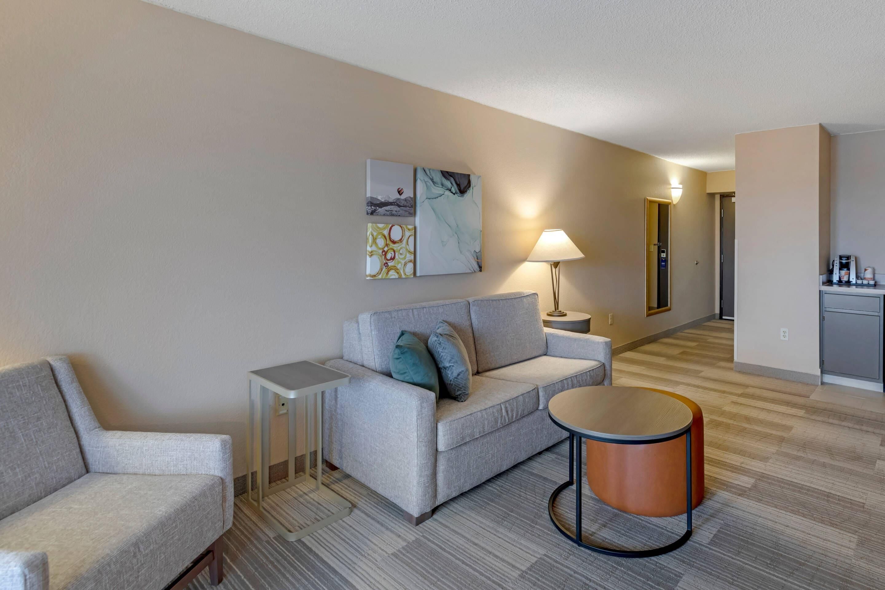 A modern hotel room with a grey sofa, a small table, a lamp, and a painting on the wall.