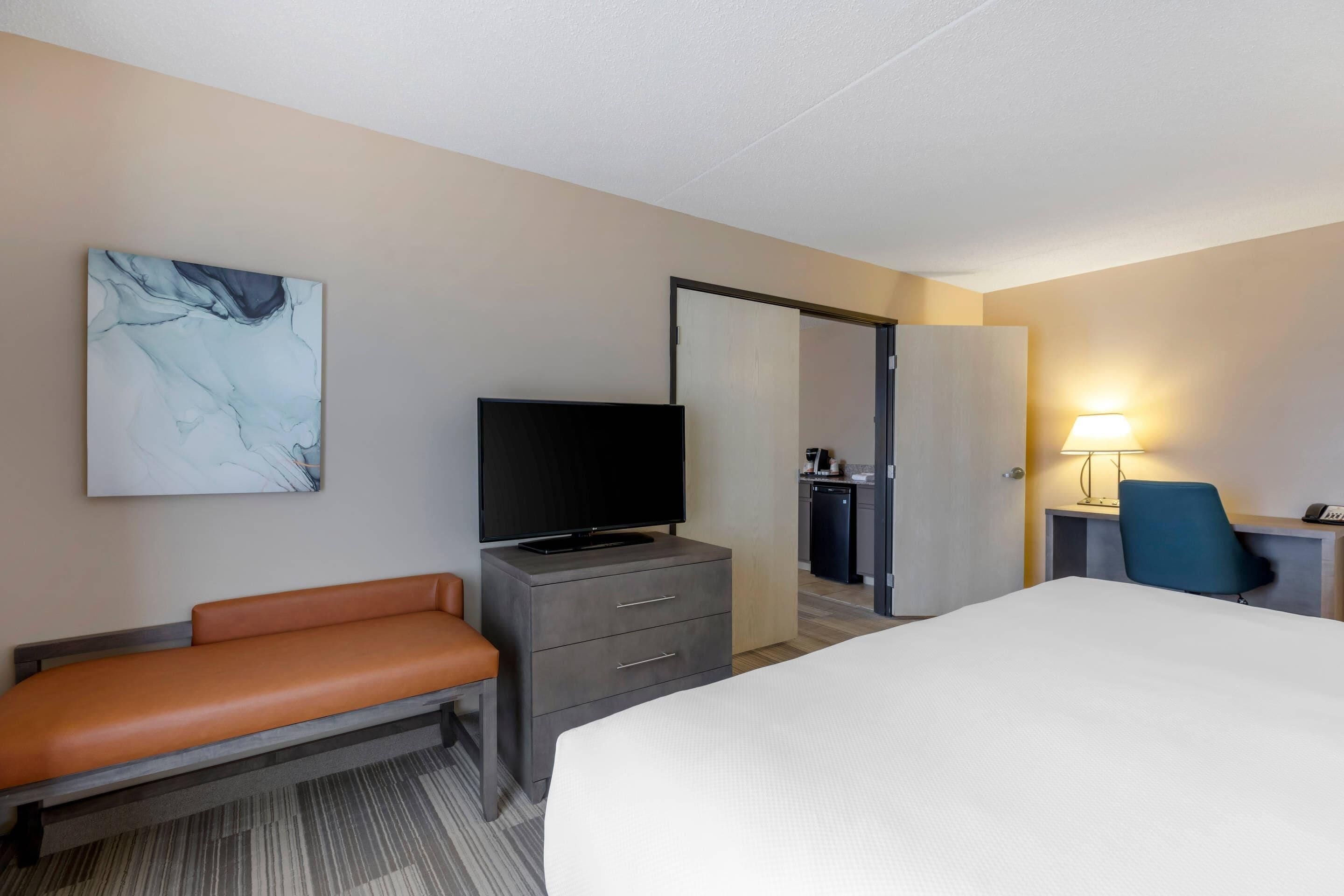 Quarto de hotel com cama, televisão em uma cômoda, escrivaninha com luminária, cadeira e banheiro visível através de uma porta aberta.