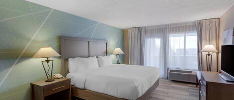 A hotel room with a large bed, two bedside tables with lamps, a TV on a wooden dresser, and a large window with curtains.
