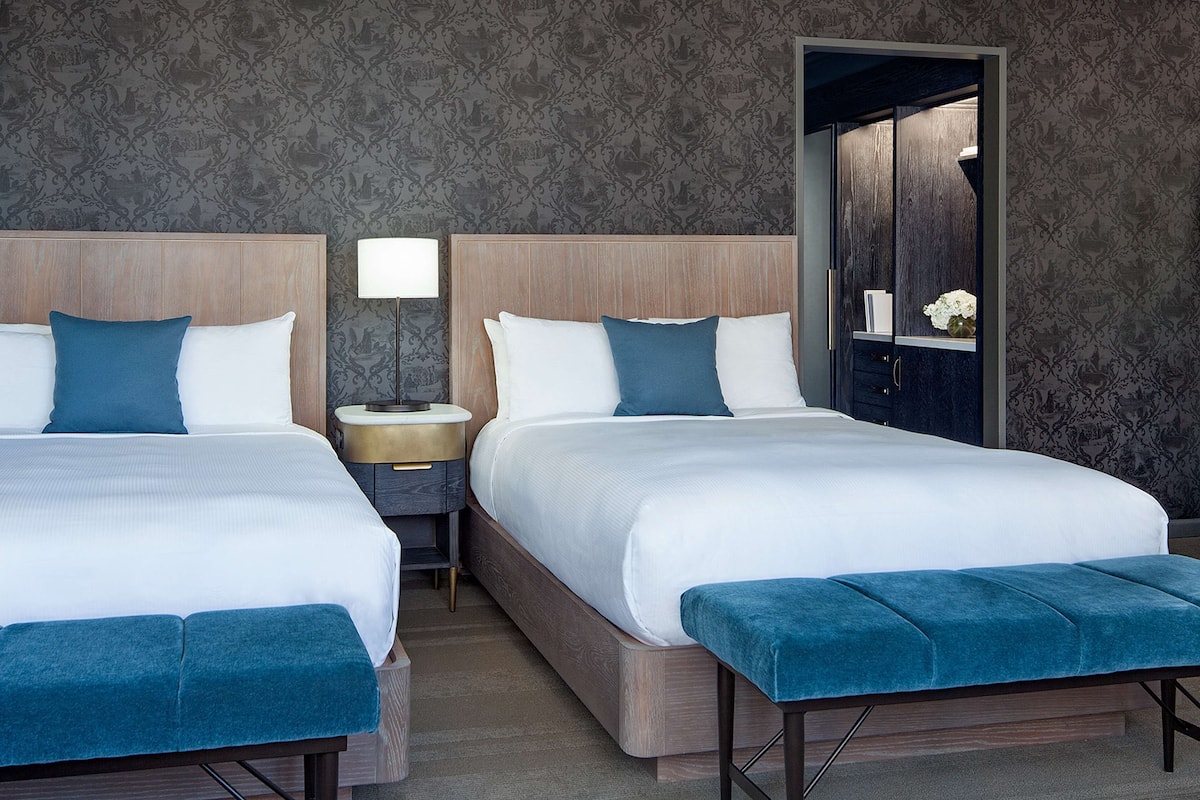 Hotellihuone, jossa on kaksi sänkyä, yöpöytä lampun kanssa, penkki ja peili.
