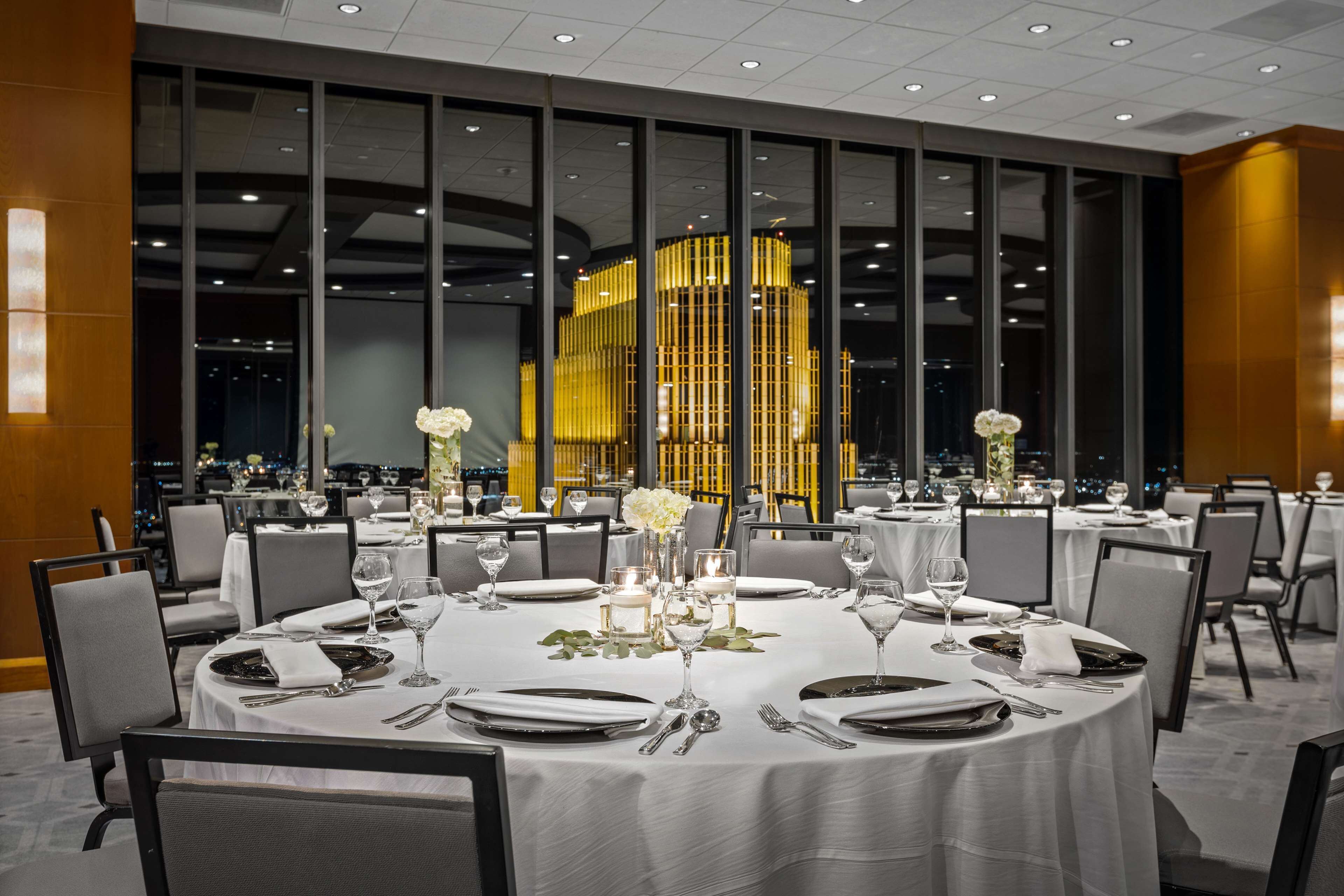 A restaurant with round tables set for dining, large windows, and a view of the city at night.