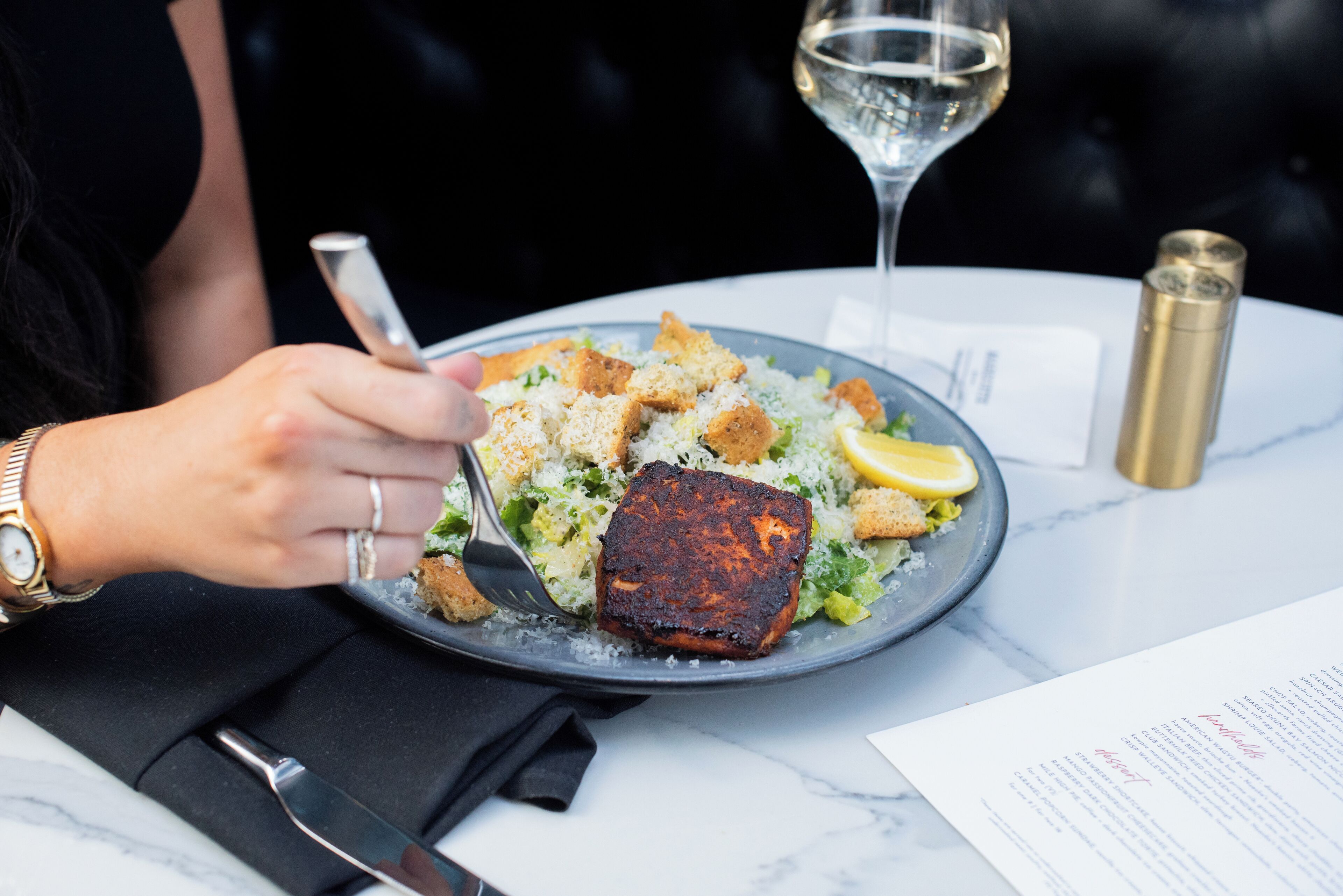 A person dining at a table with a plate of food, a glass of white wine, and a menu.