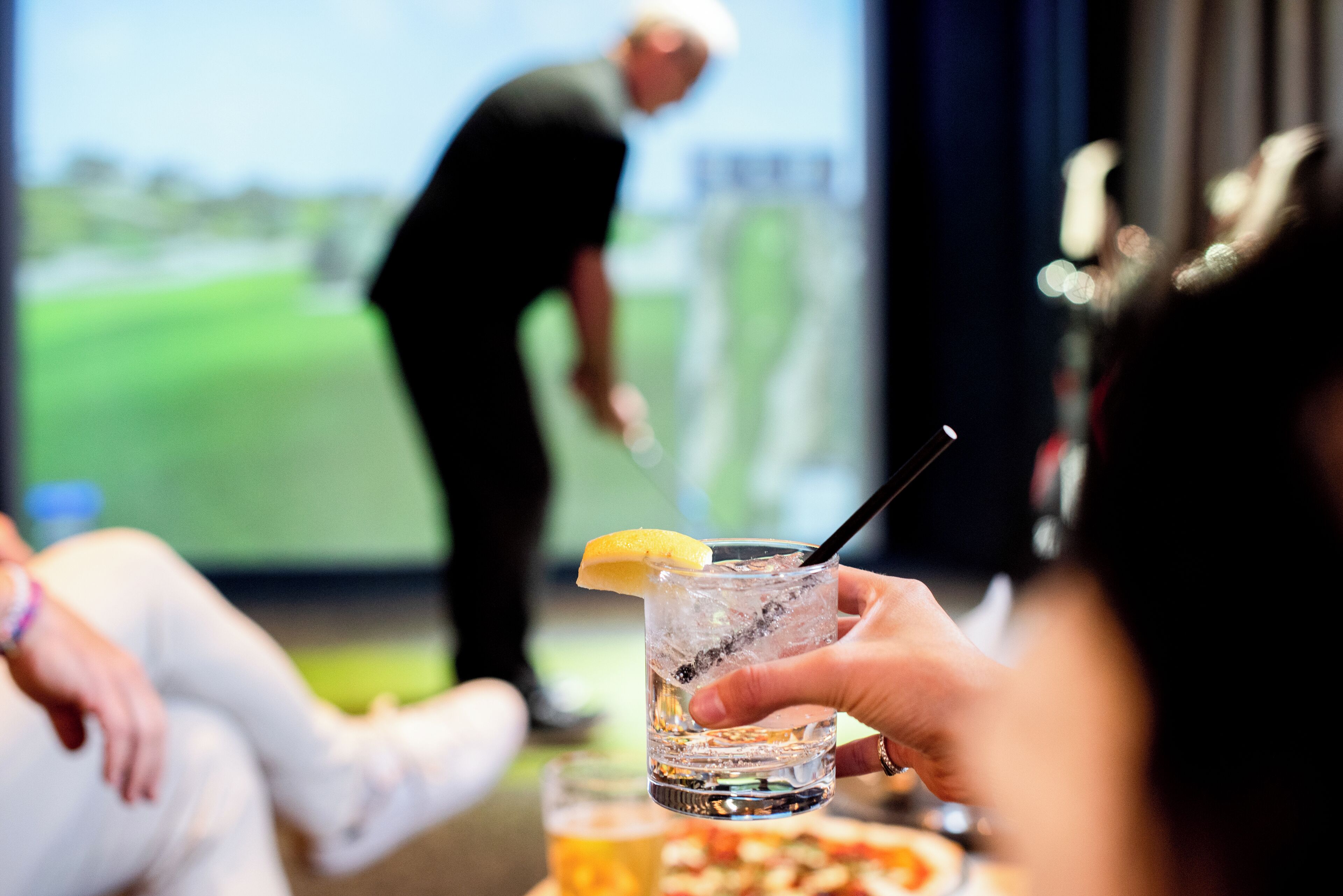 En person holder et glas med et citronskive og et sugerør, mens en golfbane ses i baggrunden.
