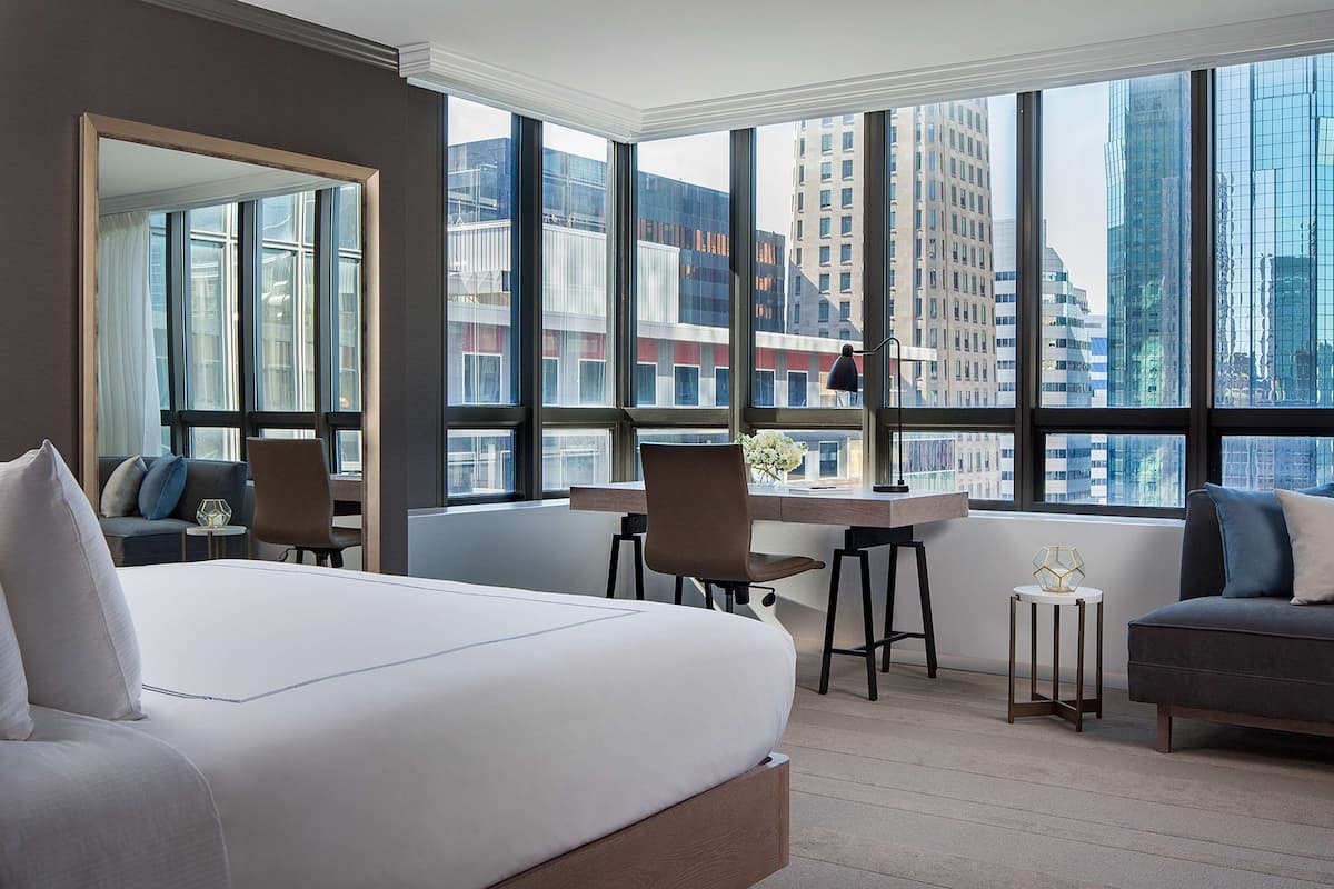 Moderni hotellihuone, jossa on suuri sänky, työpöytä ja suuret ikkunat, joista avautuu näkymä kaupungin rakennuksiin.