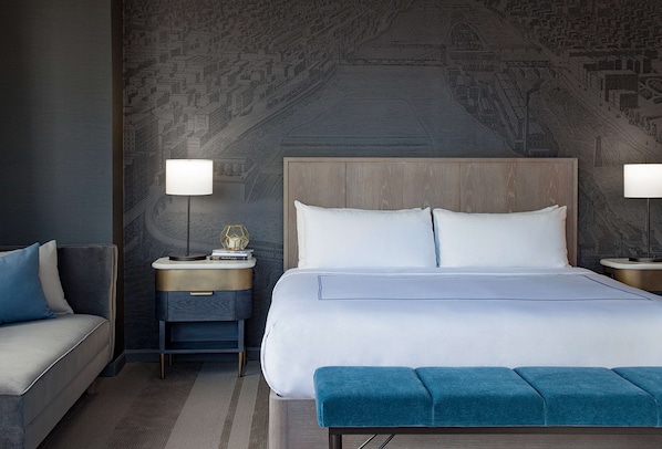 A modern hotel room with a large bed, a nightstand with a lamp, a sofa, and a patterned wall.