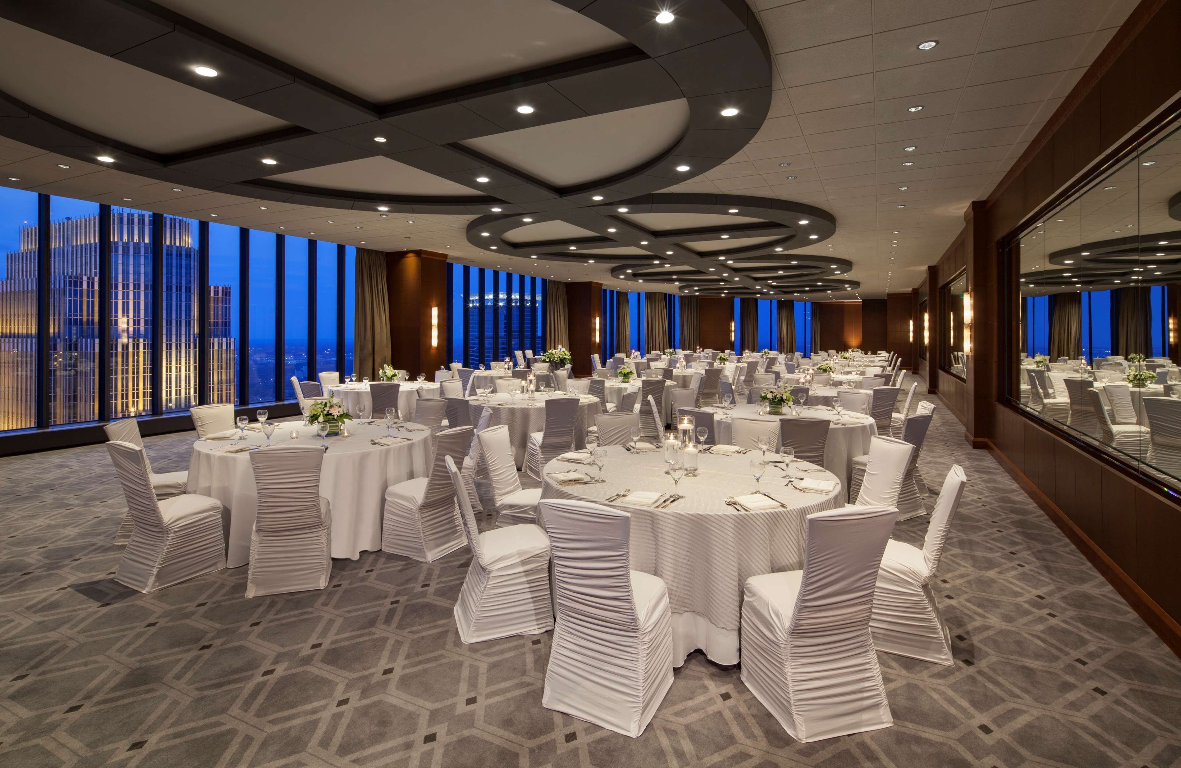 A large conference room with multiple round tables, white chairs, and a city view through large windows.
