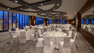 A large conference room with multiple round tables, white chairs, and a city view through large windows.
