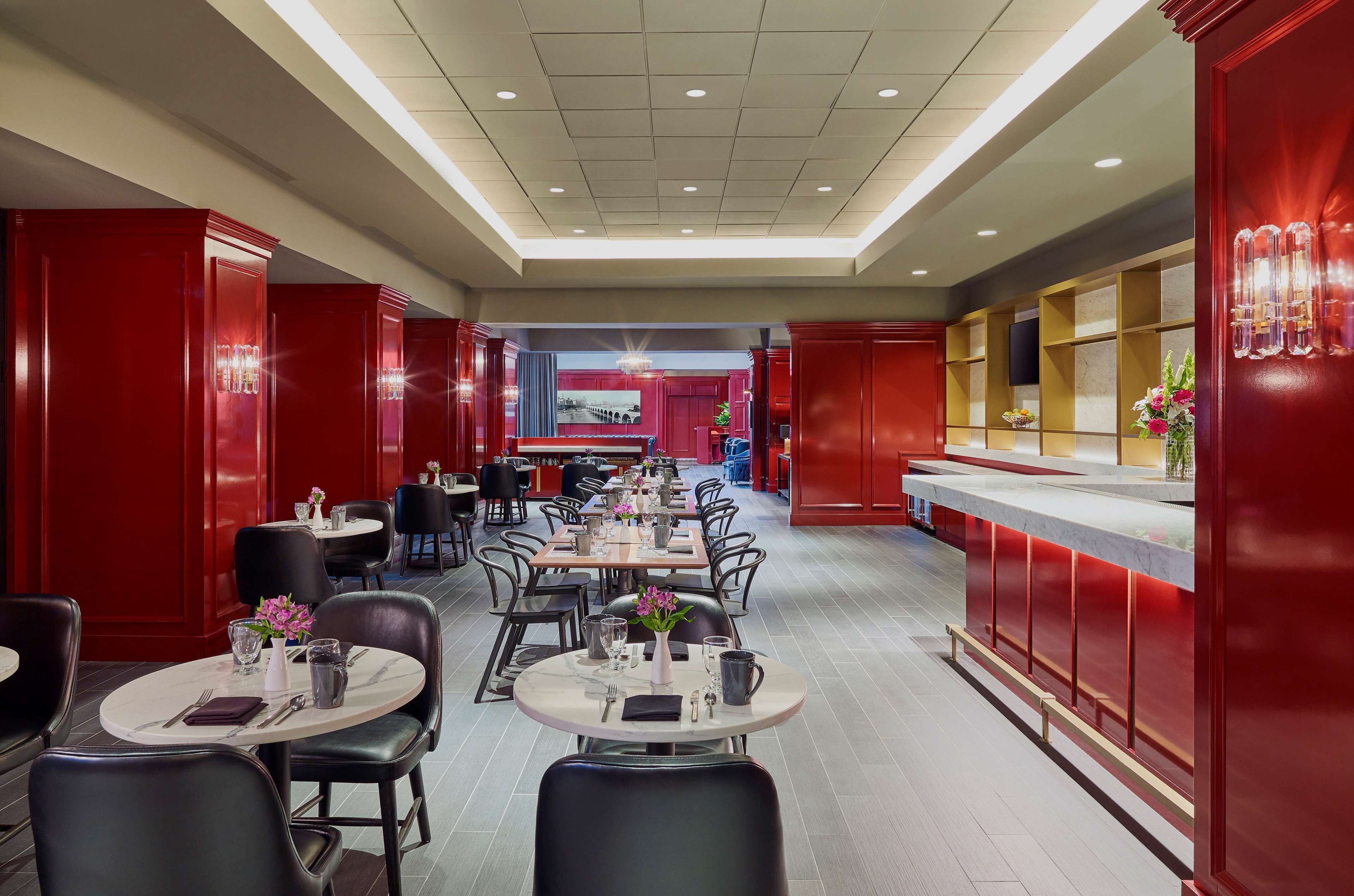 Un restaurante con varias mesas y sillas, una barra con fondo rojo y un diseño moderno en el techo.