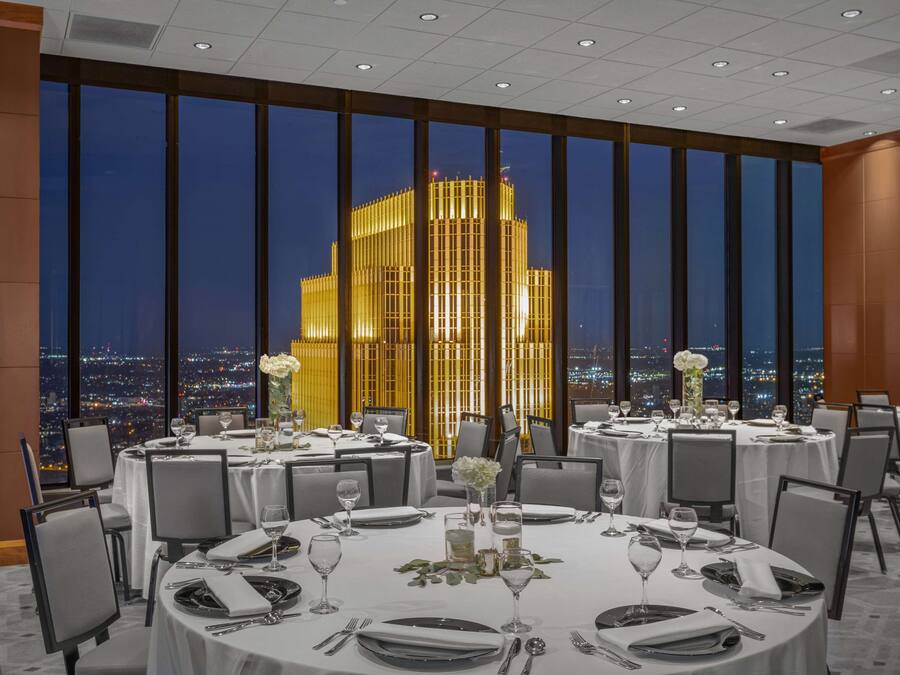 A dining area with round tables set for a meal, large windows offering a view of a cityscape at night, and a modern building illuminated in the background.