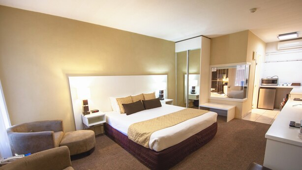 Quarto de hotel com cama grande, mesa de cabeceira, cadeira, escrivaninha e cozinha compacta.