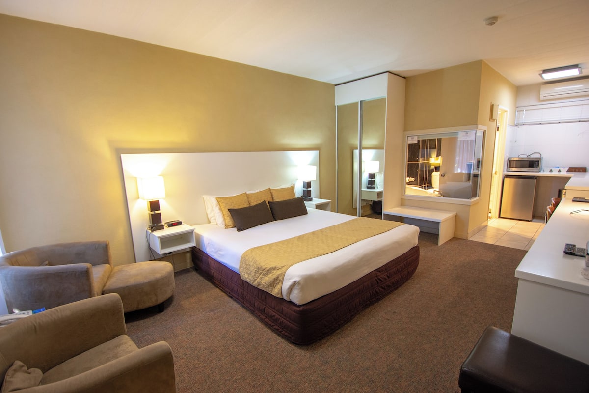 A hotel room with a large bed, a sitting area with a chair and ottoman, a small desk, and a kitchenette.
