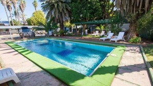 Piscina retangular com borda verde, rodeada por um terraço com cadeiras de sol e uma área coberta para relaxar.