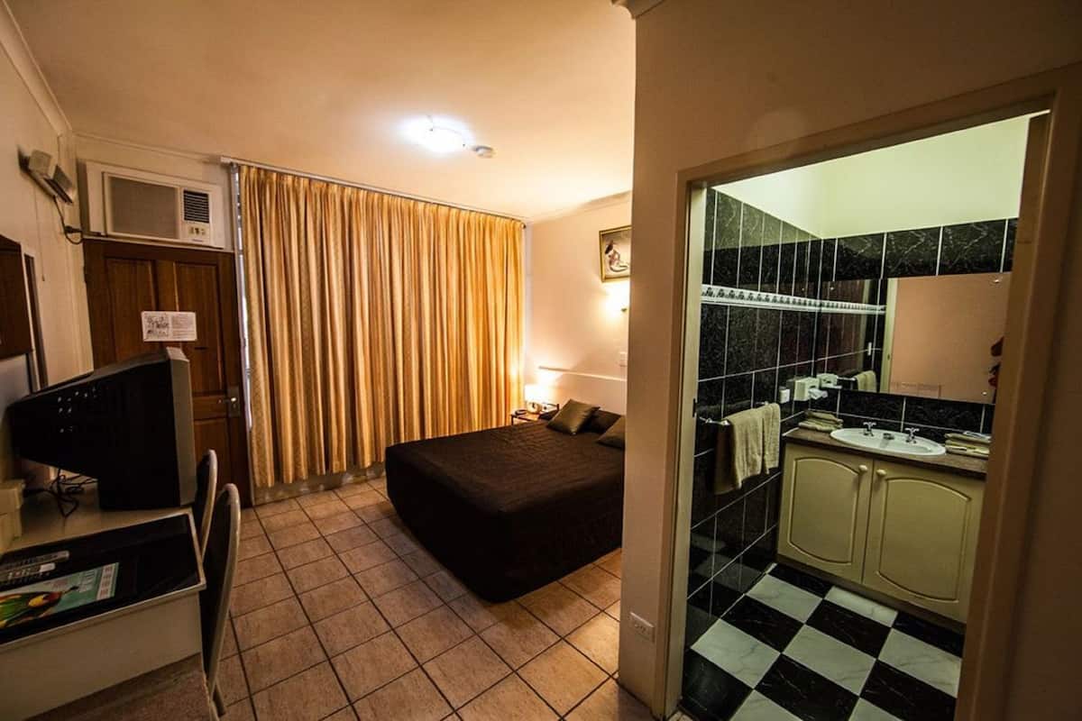 A hotel room with a bed, a desk with a computer, a window with curtains, a bathroom with a sink and mirror, and a tiled floor.