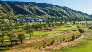 A golf course with a mountainous backdrop, a few scattered trees, and a distant building complex.