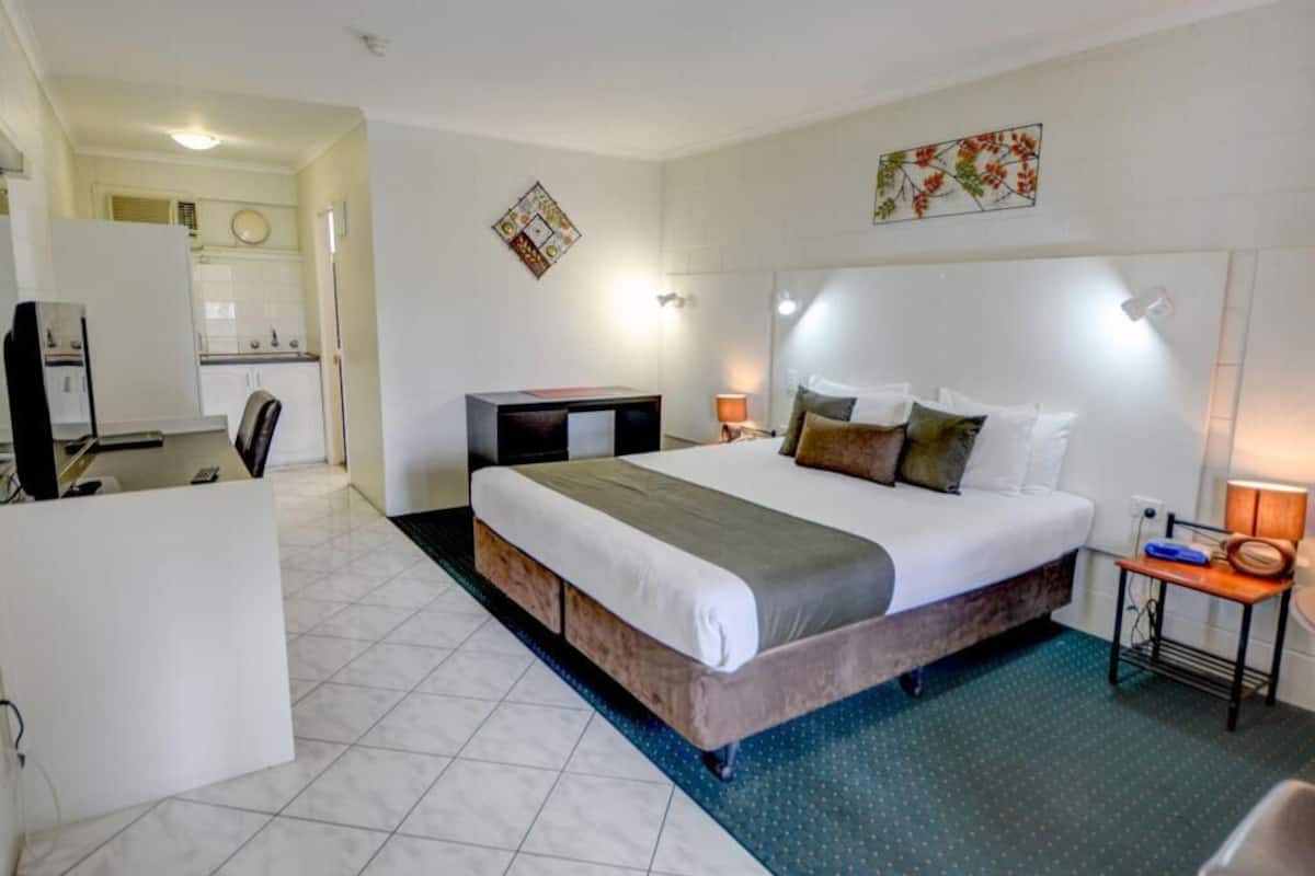 A hotel room with a bed, bedside table, lamp, and a view of the kitchen area.