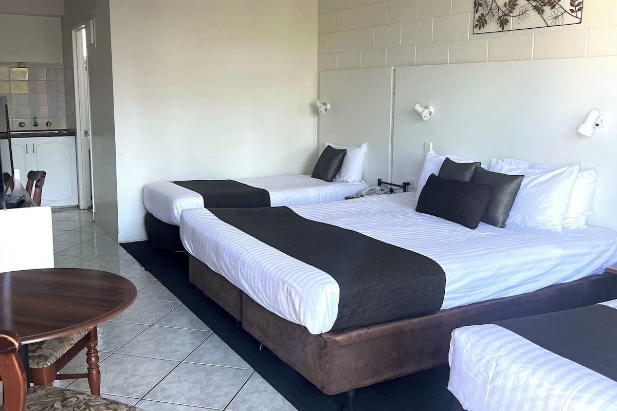 A hotel room with a large bed, a small table, and a chair. There is a kitchenette in the background.