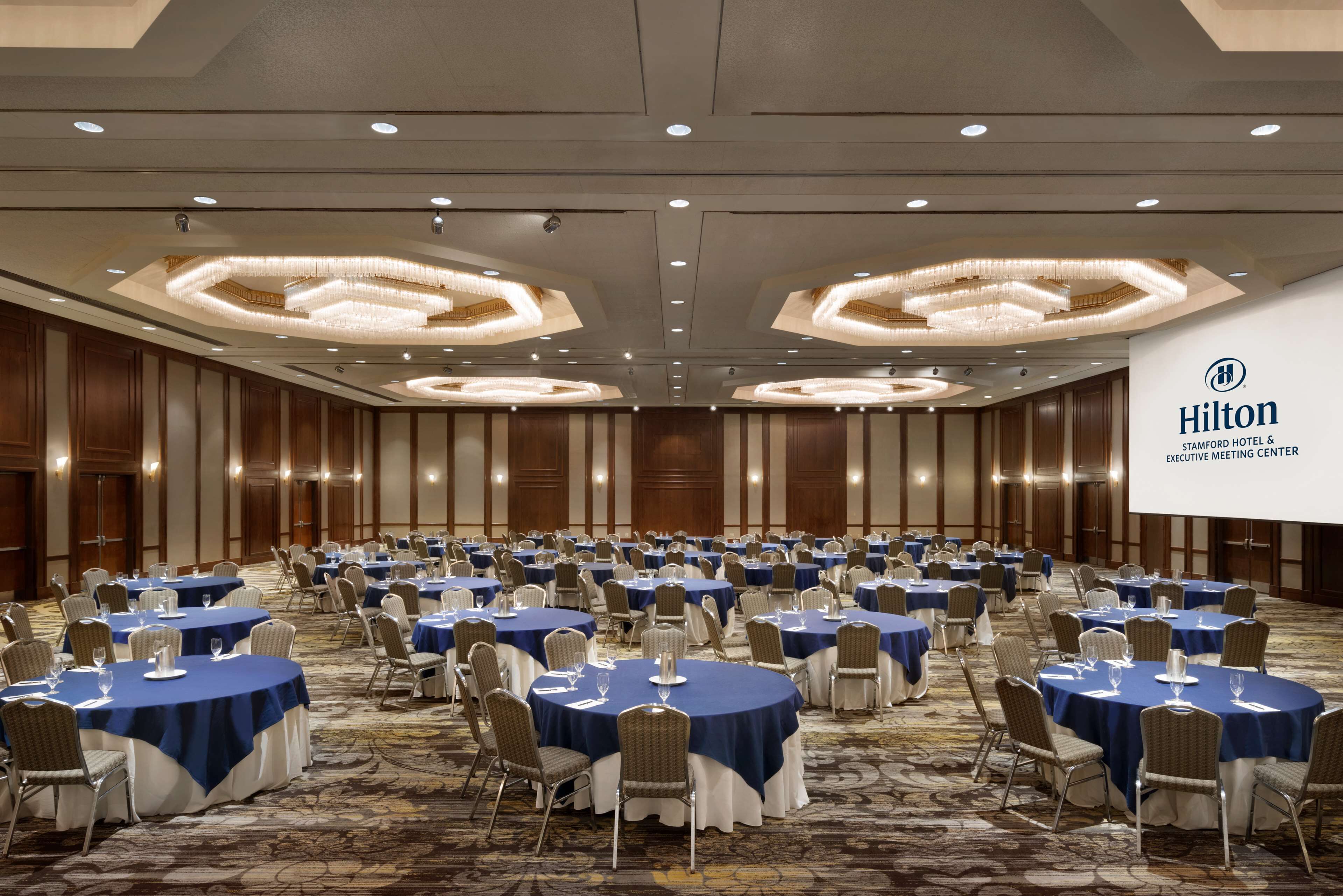A large, well-lit conference room with round tables covered in blue tablecloths and white chairs, featuring a prominent Hilton logo on the wall.