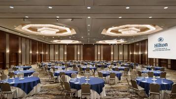 Una amplia sala de conferencias bien iluminada con mesas redondas cubiertas con manteles azules y sillas blancas, con un llamativo logotipo de Hilton en la pared.