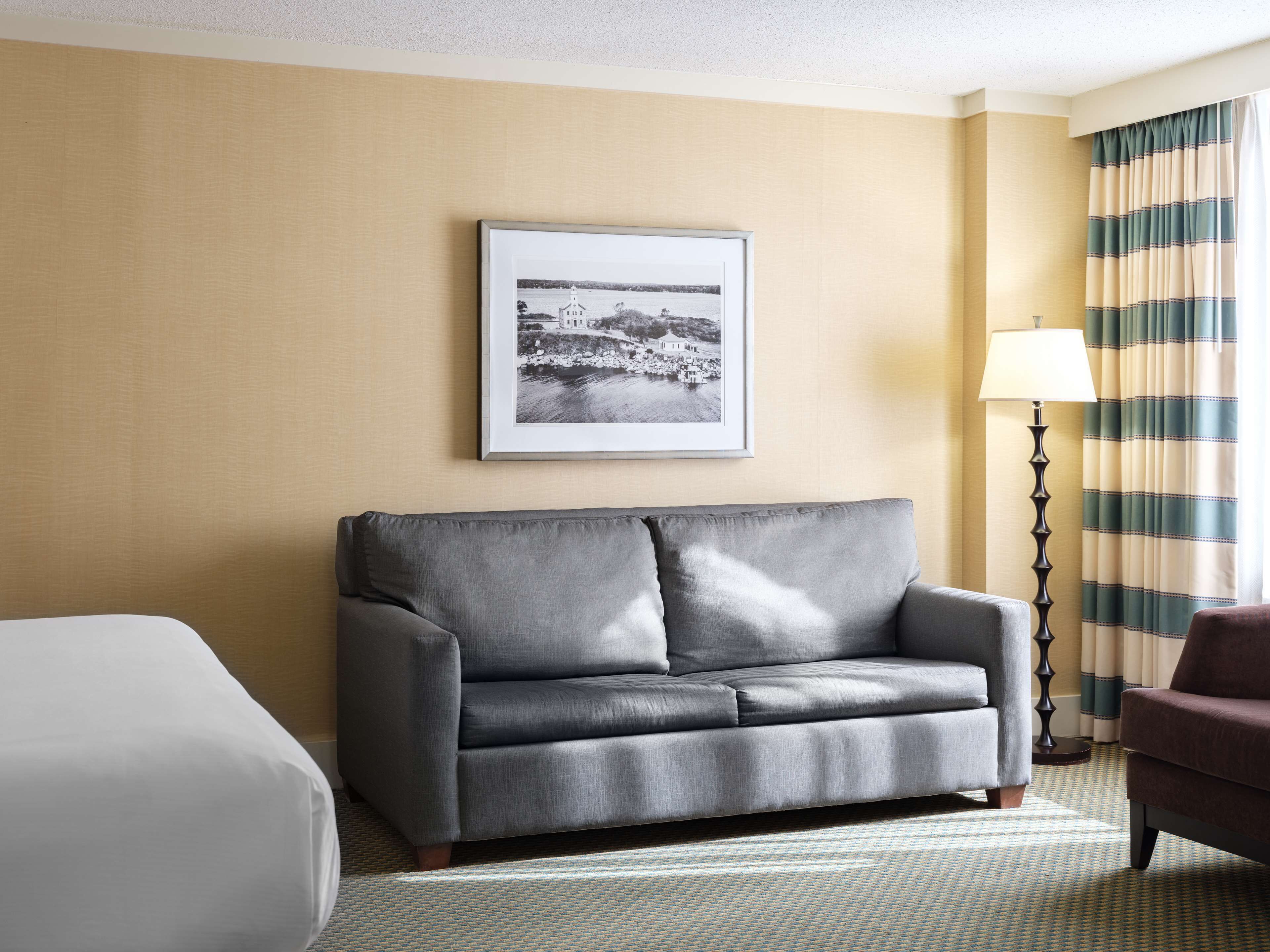 Quarto de hotel com um sofá cinzento, uma fotografia a preto e branco emoldurada na parede, uma luminária e cortinas listradas.